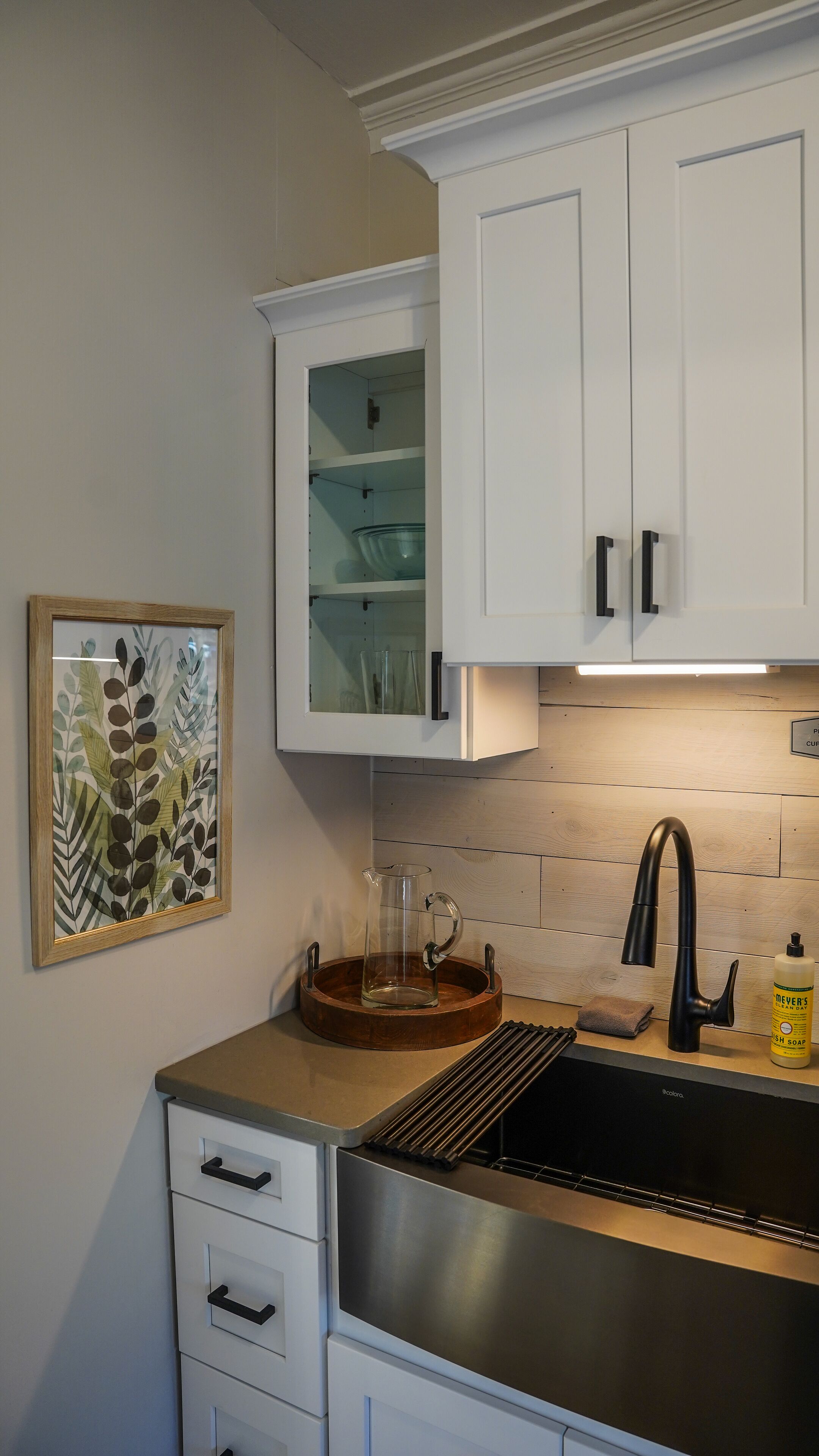 Una cocina con armarios blancos, un lavamanos de acero inoxidable, una cafetera y una botella de detergente.