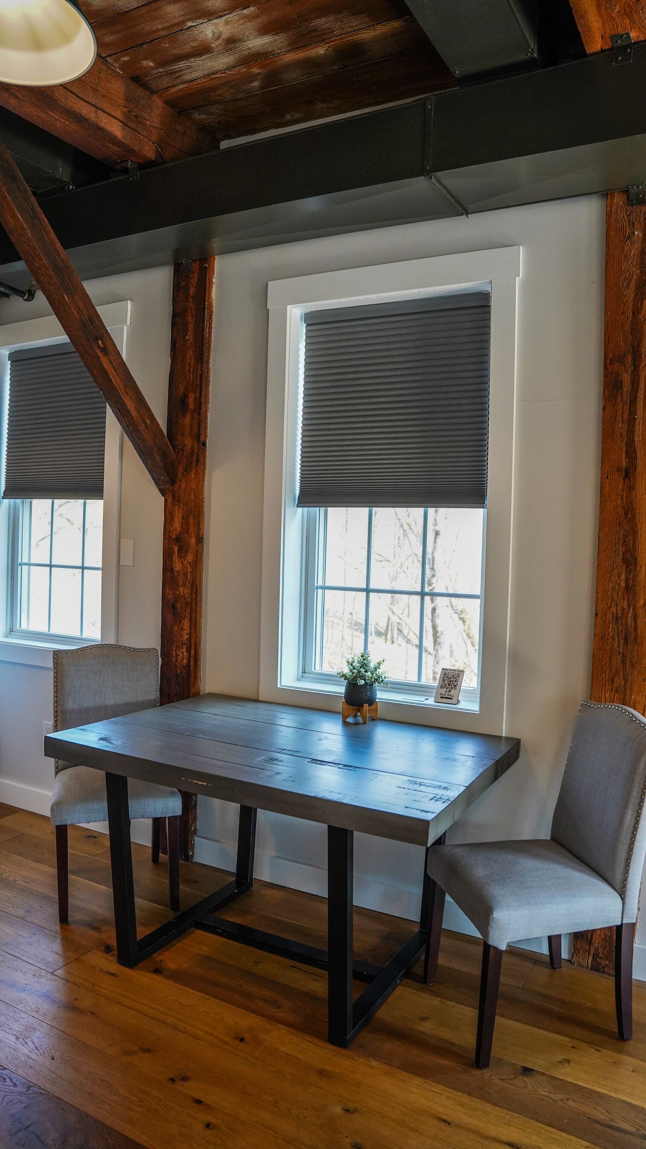 Un comedor con mesa y sillas de madera, ventanas amplias con persianas y vigas de madera a la vista en el techo.