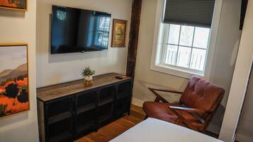 A room with a bed, a TV mounted on the wall, a window with blinds, a small plant on a wooden console, and a chair.