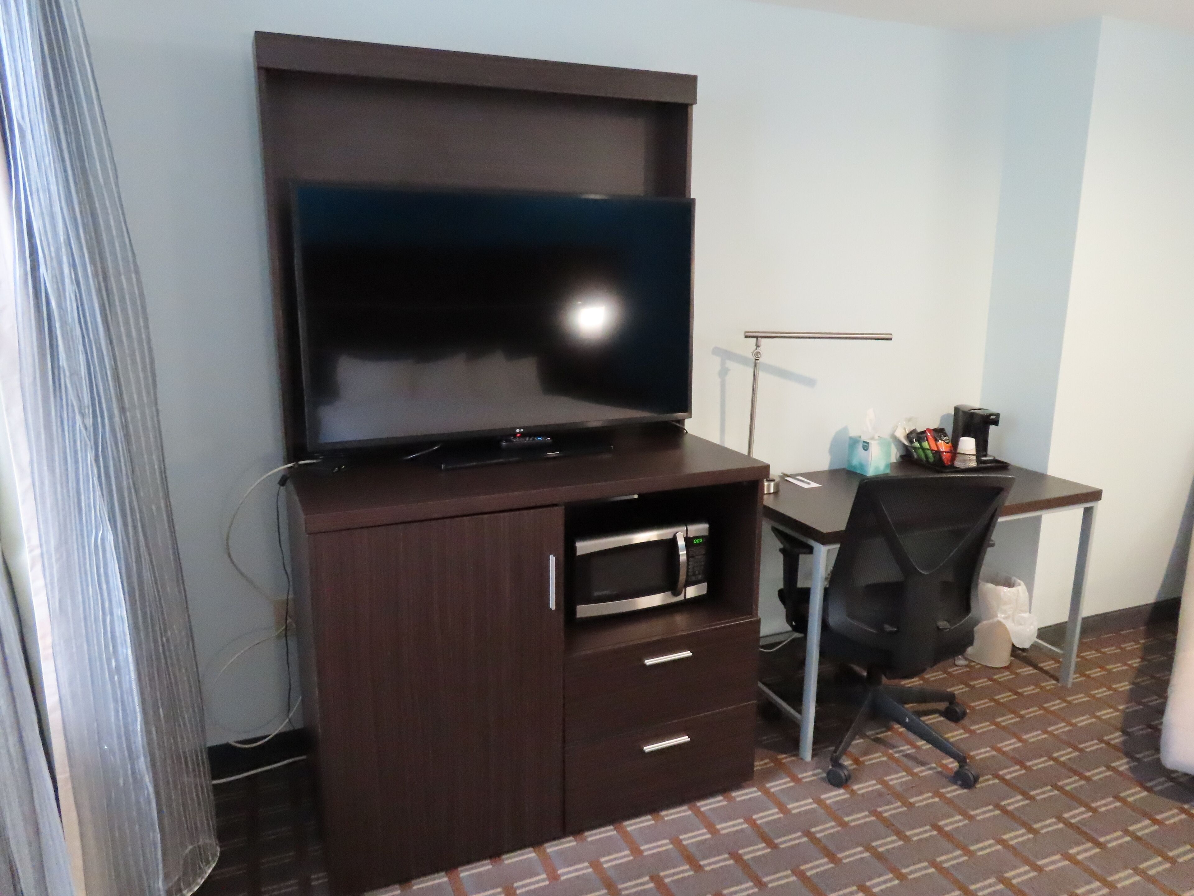 A room with a large flat-screen TV on a wooden cabinet, a desk with a chair, and a coffee machine.
