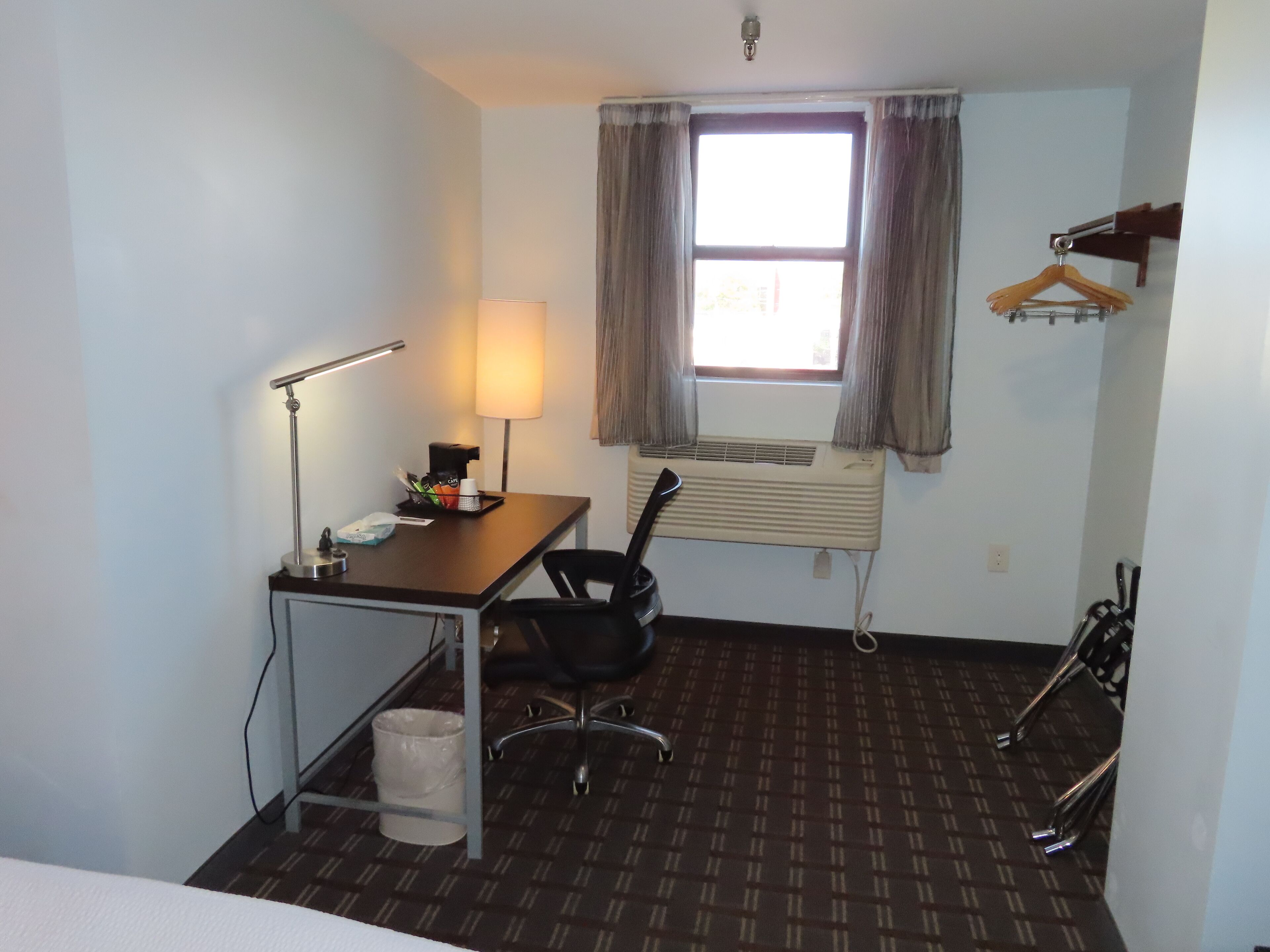Habitación de hotel con escritorio, silla, lámpara, equipo de aire acondicionado y ventana con cortinas.