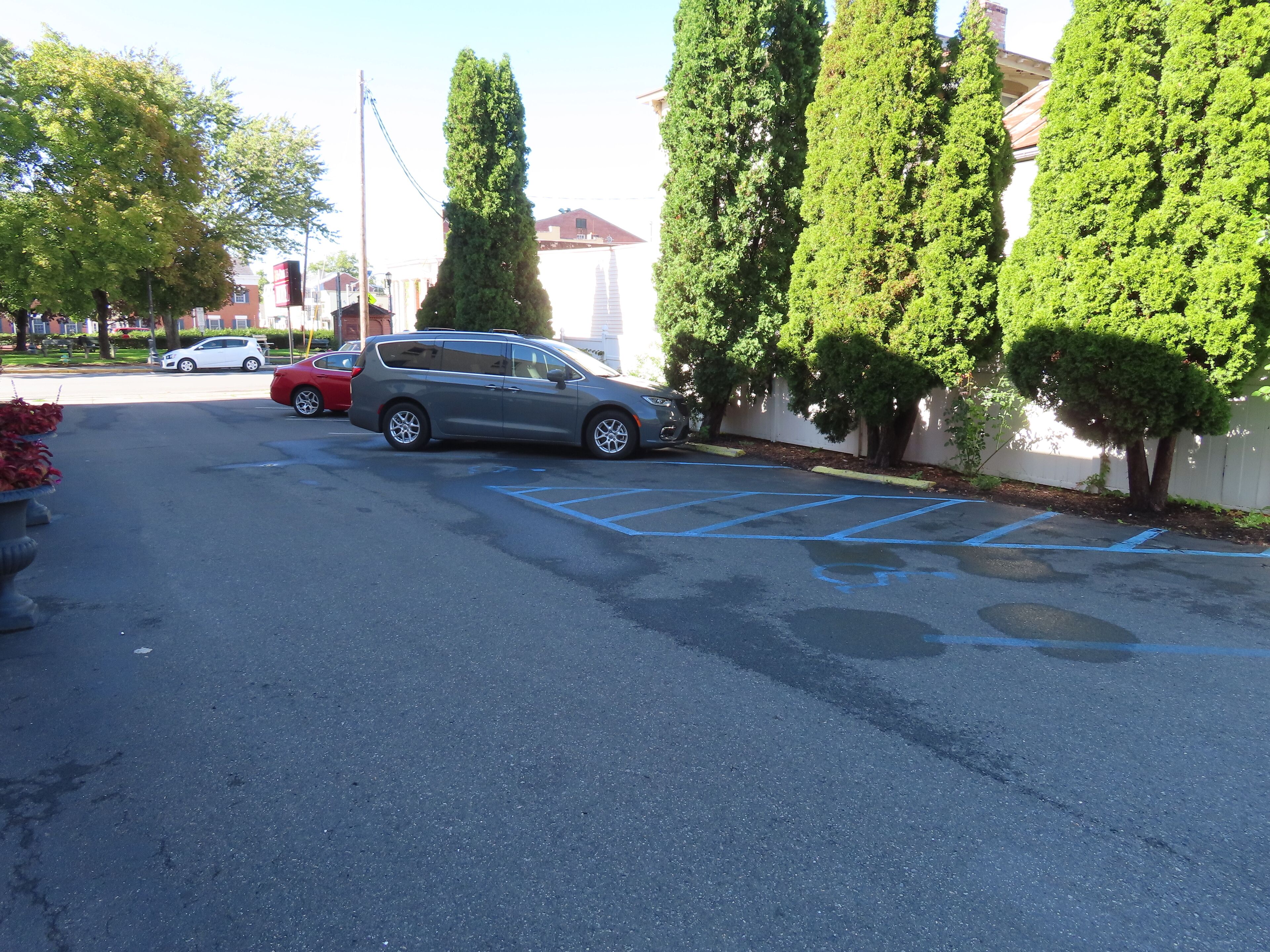 A parking lot with several cars, including a gray minivan and a red SUV. There are tall, narrow trees lining the parking area.