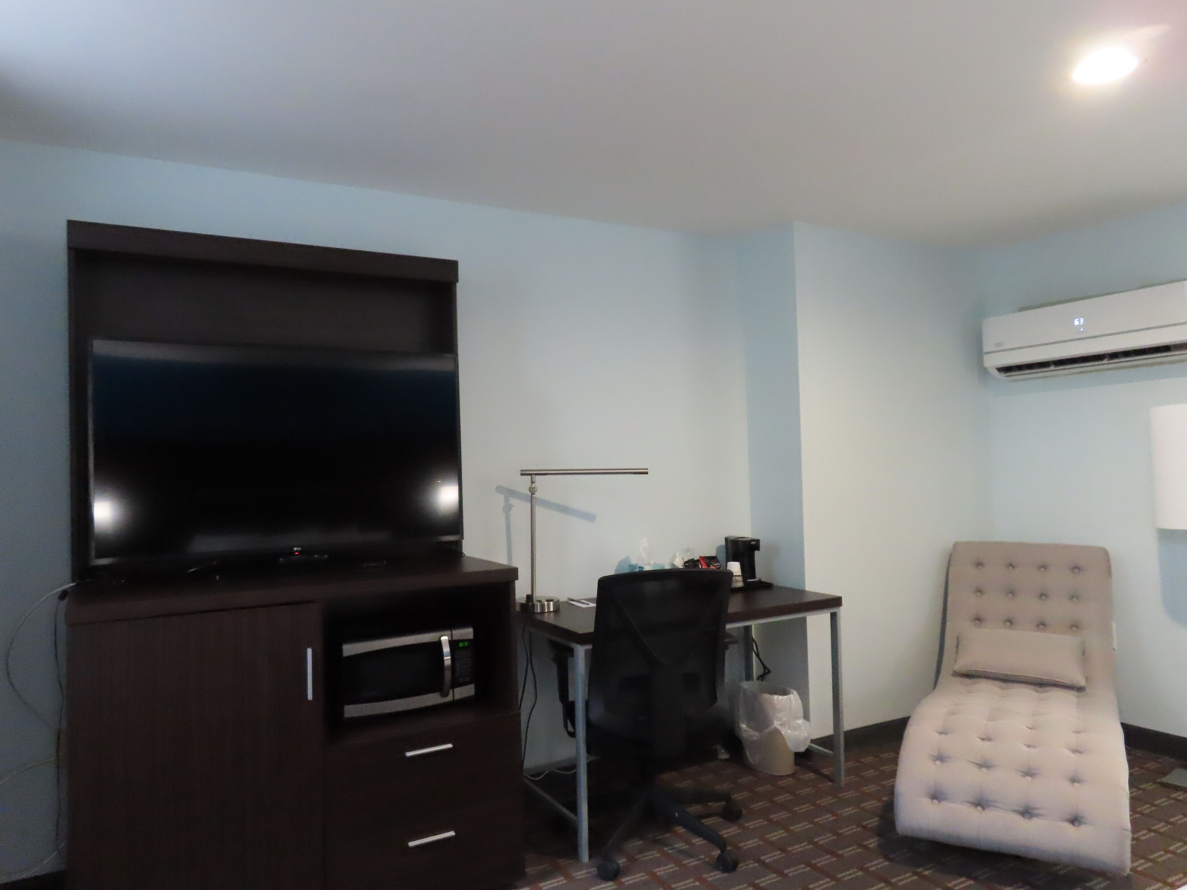 A room with a flat-screen TV on a dark wooden cabinet, a desk with a chair, and a cushioned armchair.