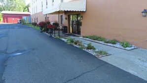 Un bâtiment à la façade beige, surmonté d’un auvent blanc au-dessus de l’entrée. Un trottoir bordé de petites plantes et une rue se trouvent devant.