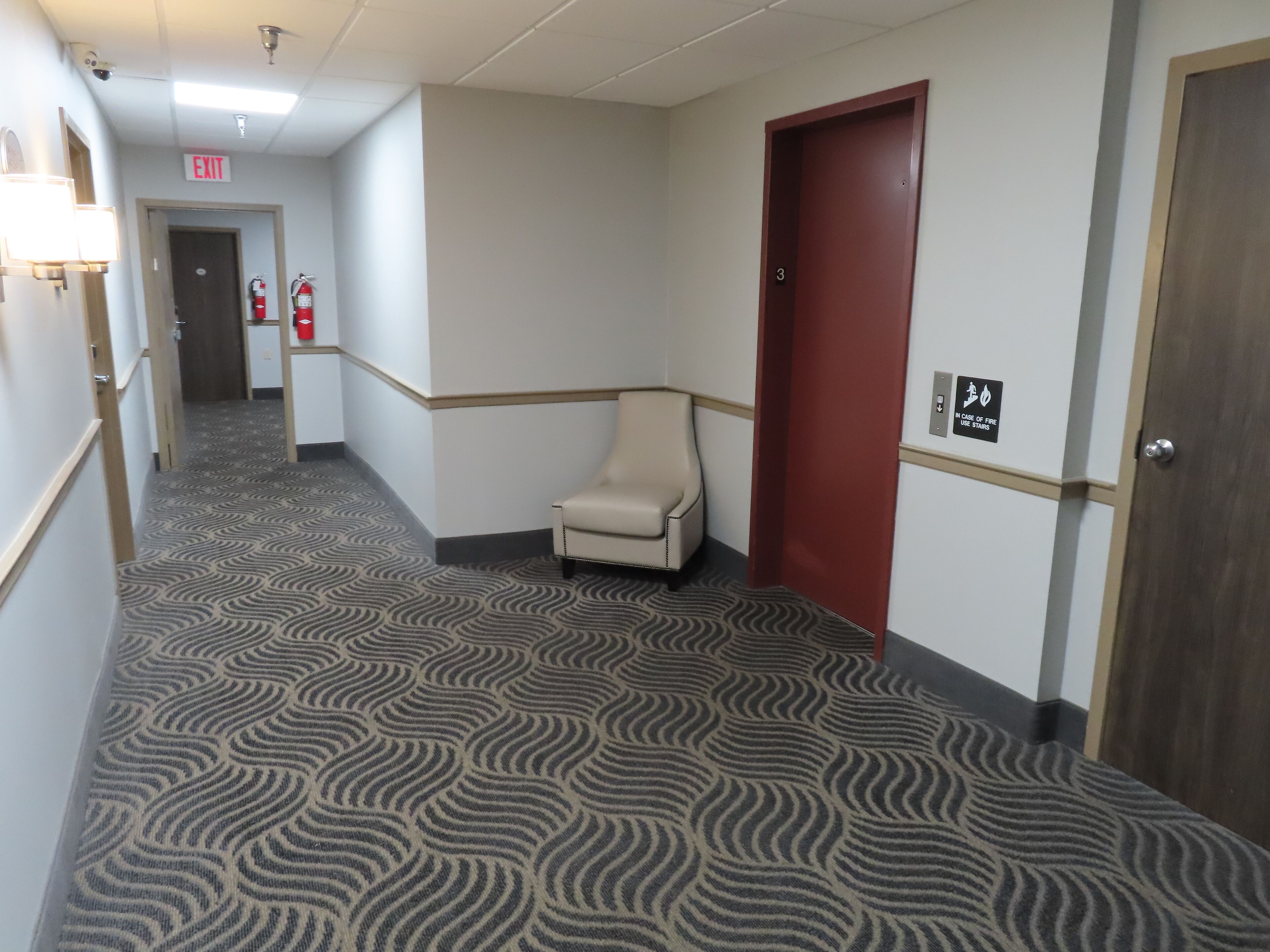 Un couloir d’hôtel avec un tapis à motifs, un extincteur, un panneau indiquant la sortie et une porte portant un symbole de sécurité incendie.