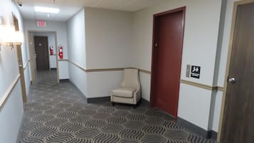 Un couloir d’hôtel avec un tapis à motifs, un extincteur, un panneau indiquant la sortie et une porte portant un symbole de sécurité incendie.