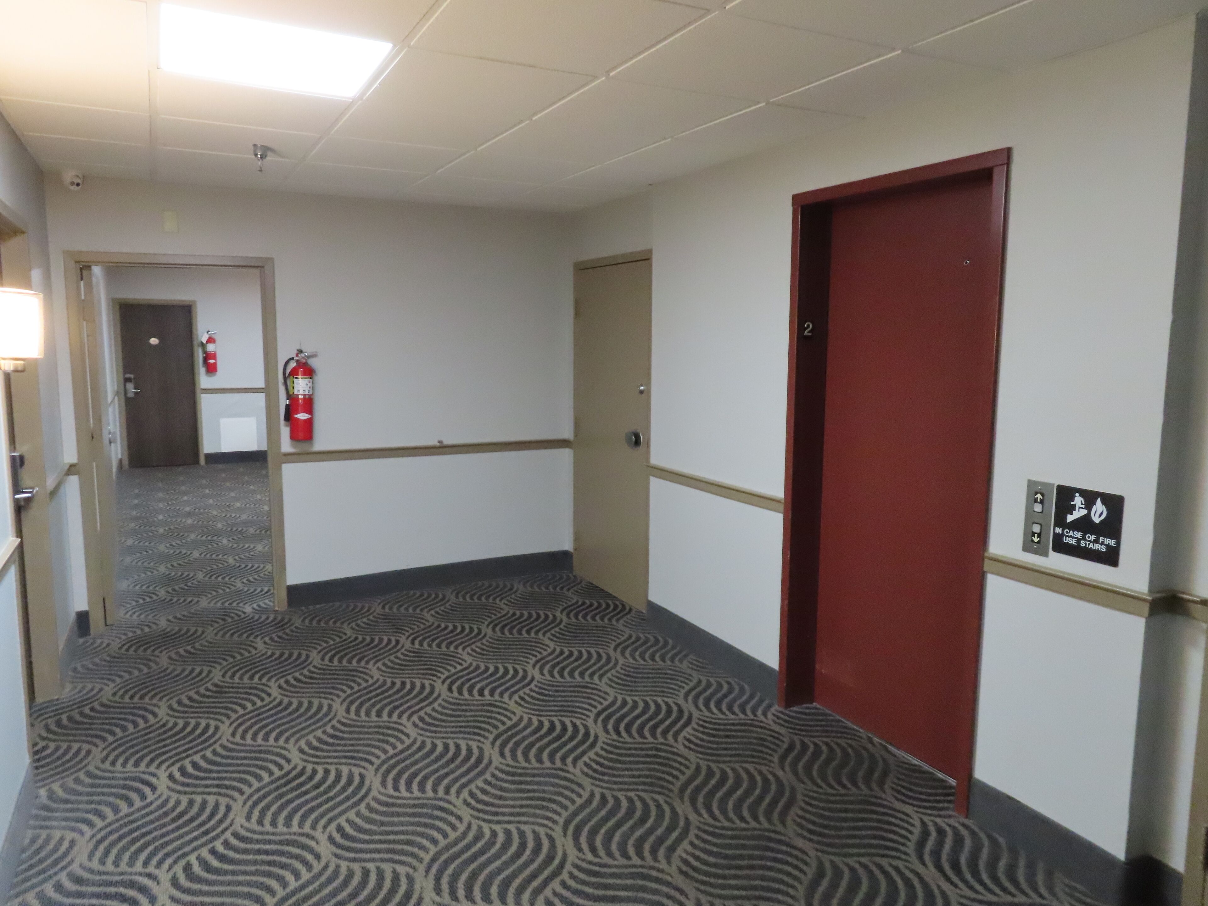 Un pasillo con alfombra estampada, una puerta roja y un extintor en la pared.