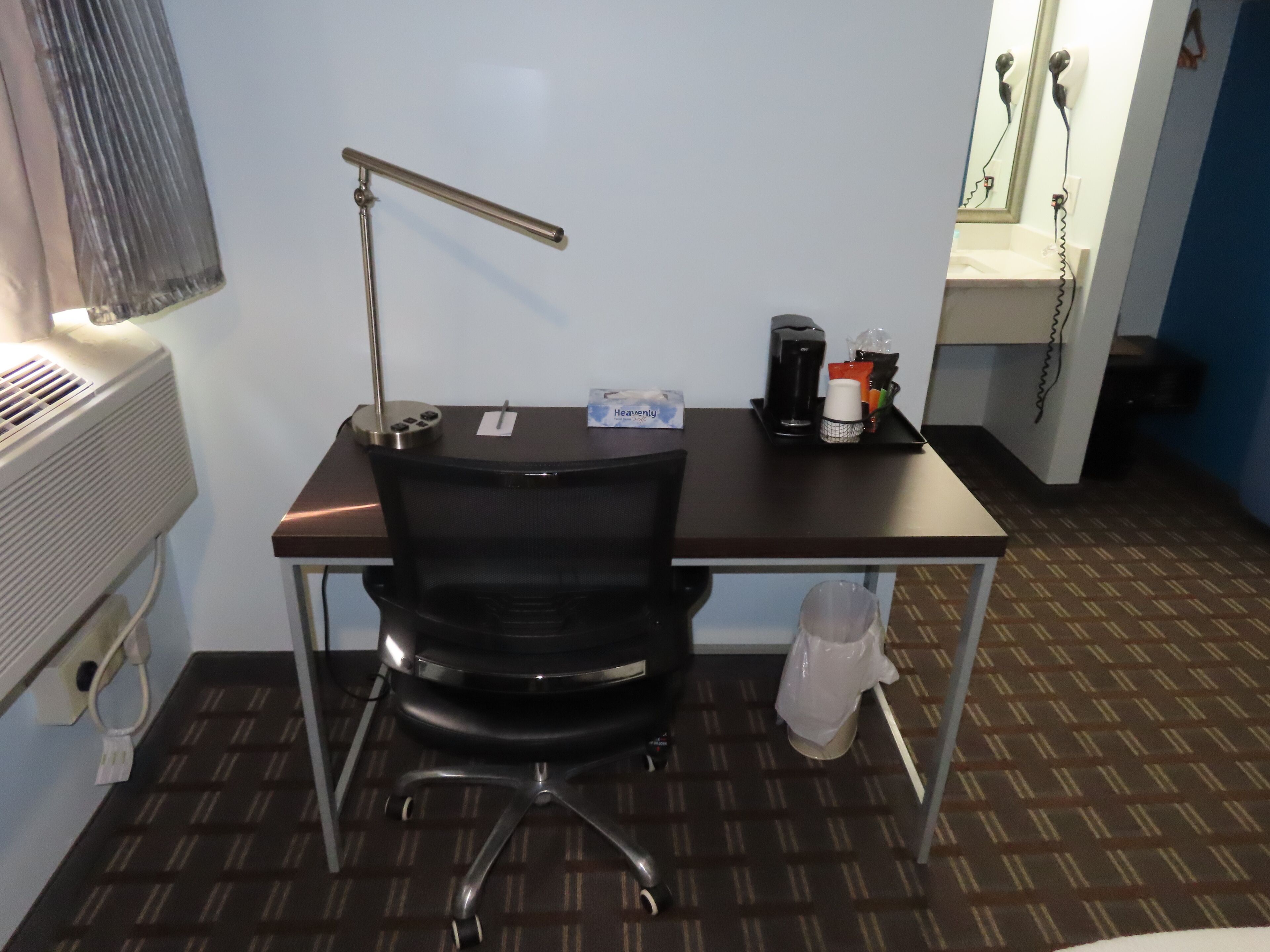 A hotel room with a desk, chair, and a small kitchenette area.
