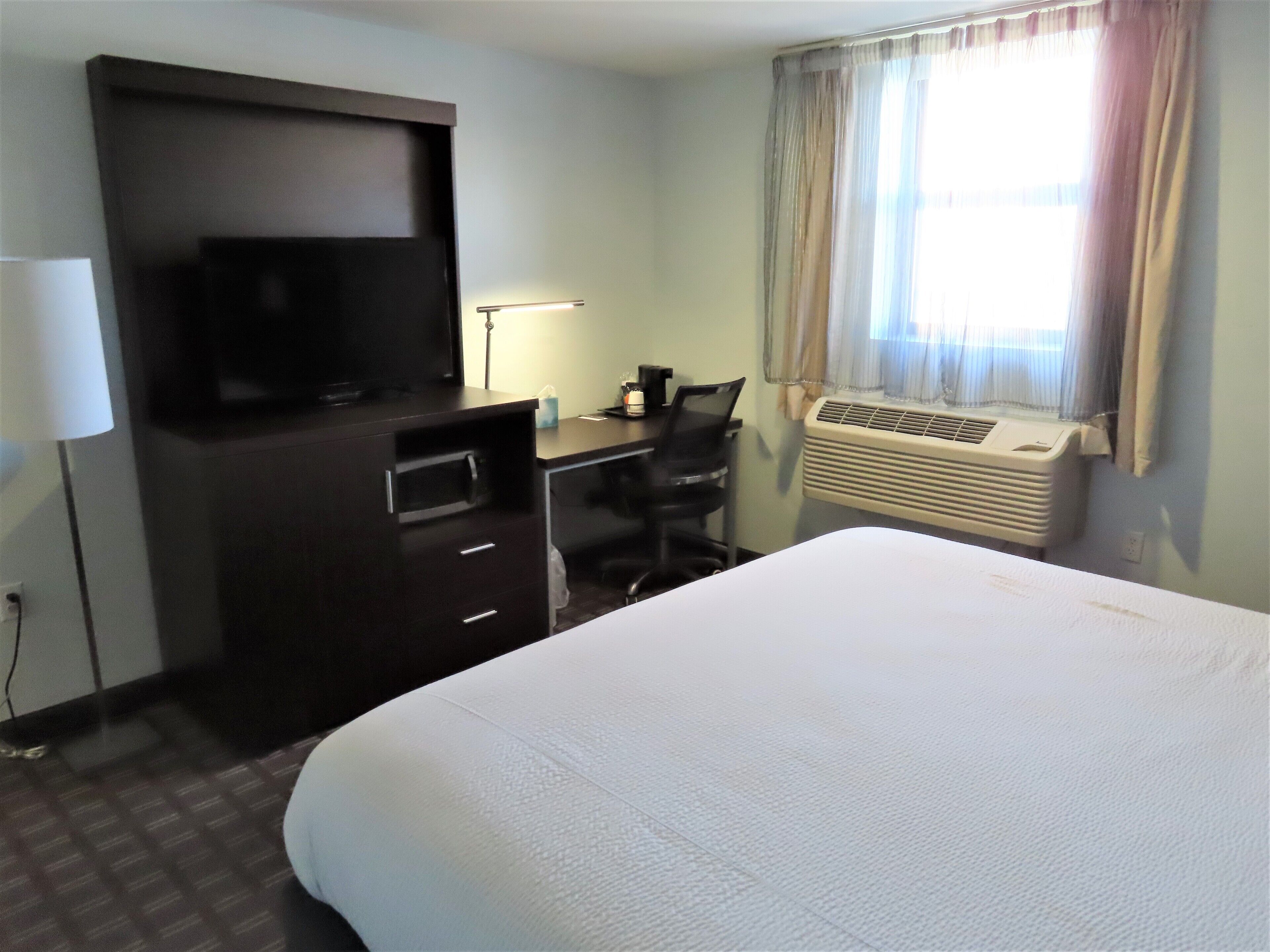 A hotel room with a bed, a desk with a chair, a television, a lamp, and an air conditioner.