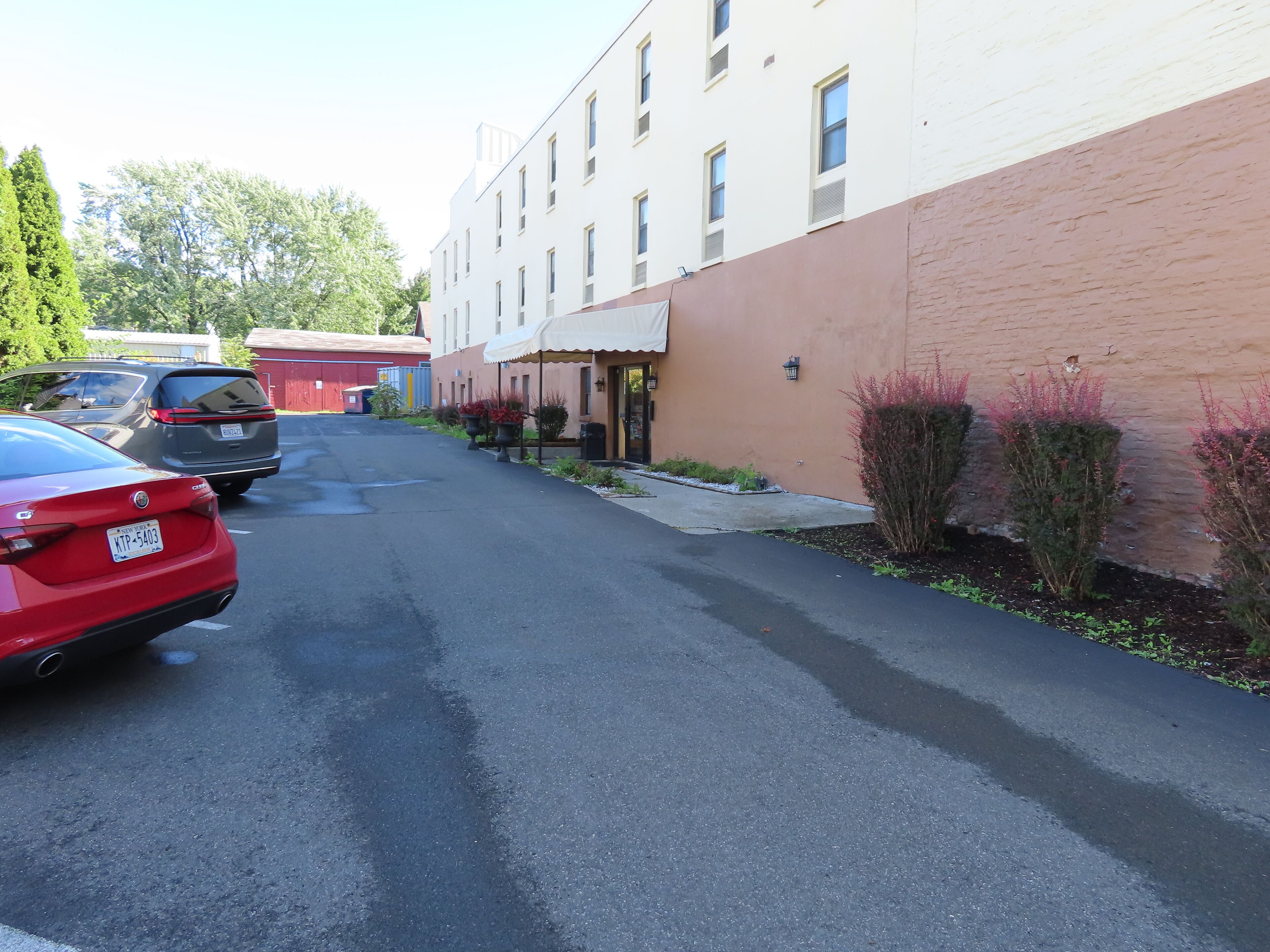 A parking lot with a red car and a gray van, a building with a beige facade, and a small garden area with shrubs and a pathway.