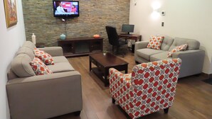 Un salon avec un mur en pierre, un téléviseur, un bureau avec un ordinateur et deux canapés ornés de coussins à motifs.