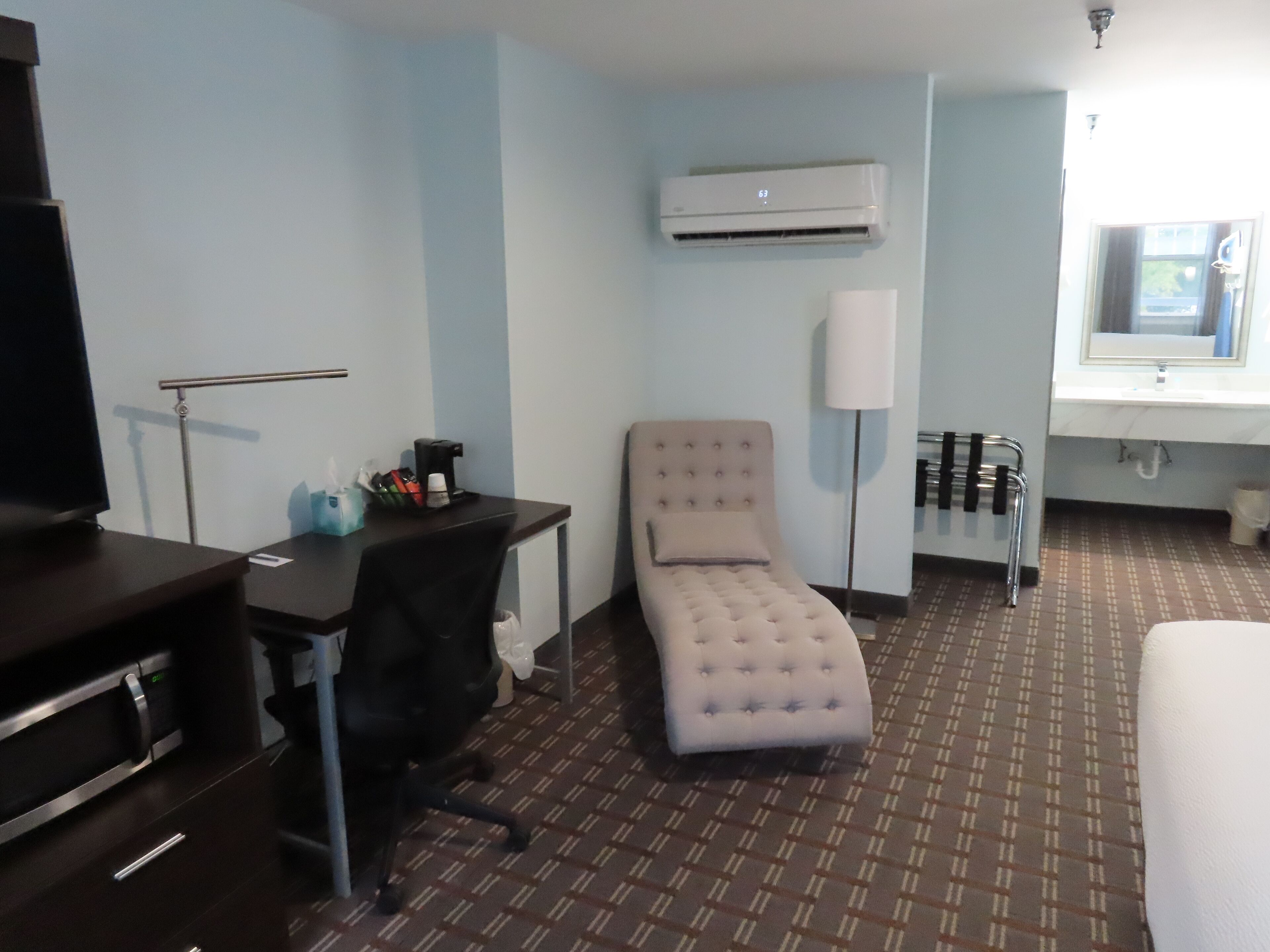 A hotel room with a bed, a desk, a chair, an air conditioning unit, a lamp, and a window.