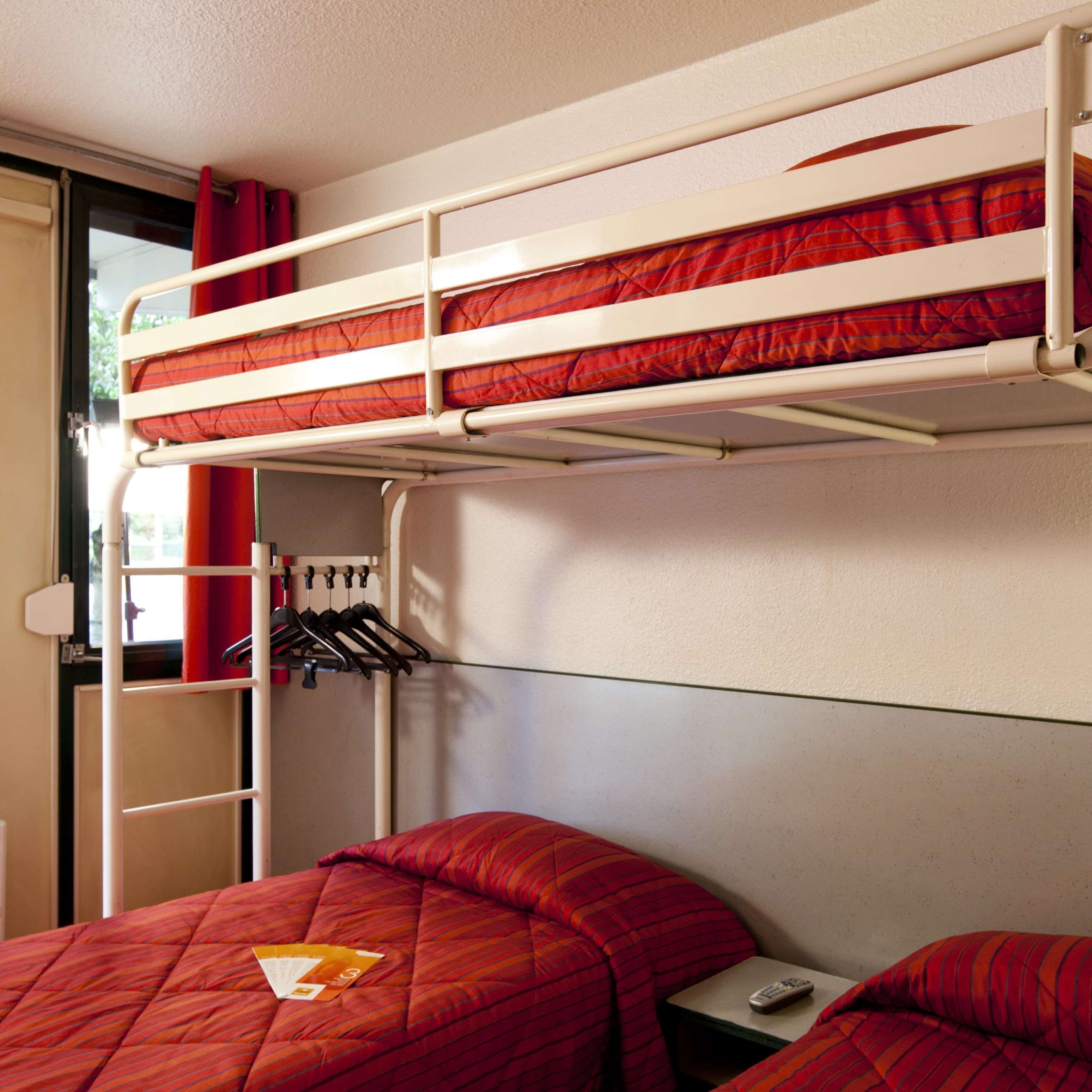 A bunk bed with a red-striped mattress, a small bedside table with a remote control, and a clothing rack with hangers.