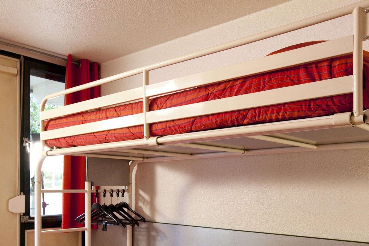 A bunk bed with a red-striped mattress, a small bedside table with a remote control, and a clothing rack with hangers.