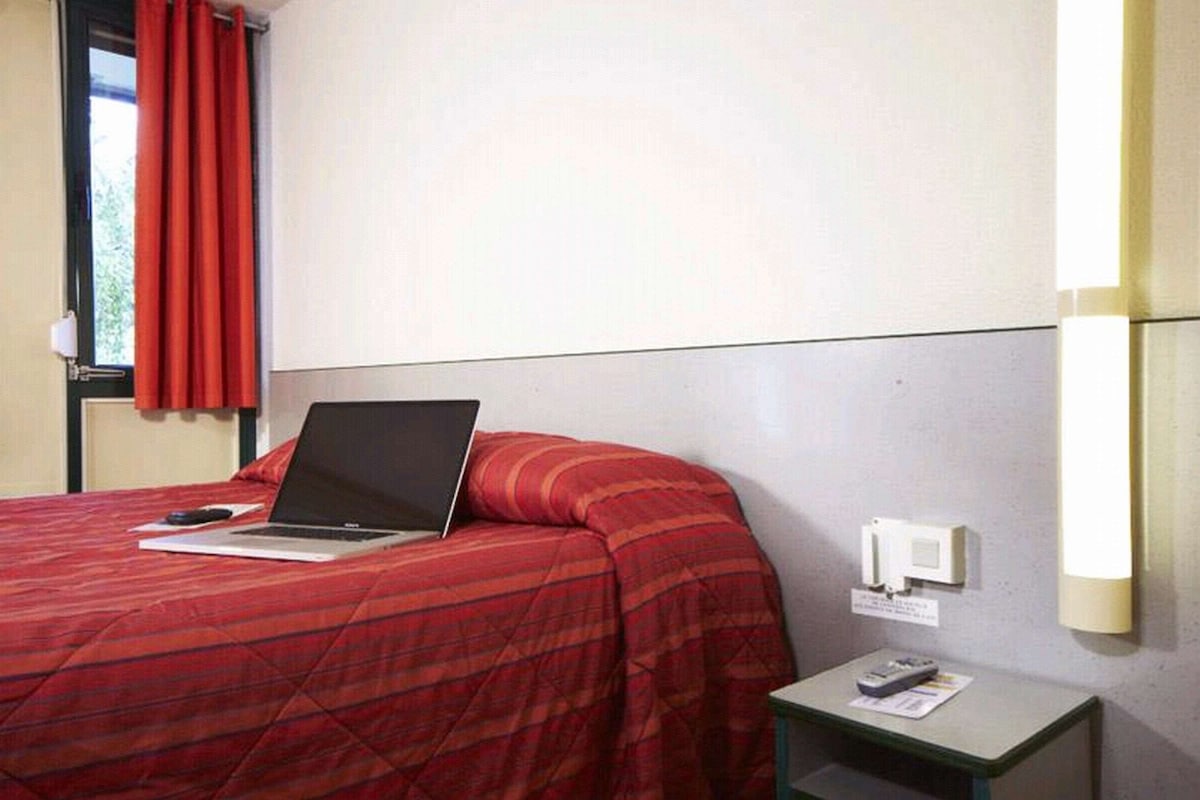 A hotel room with a bed, a laptop on the bed, a nightstand with a remote control, and a wall-mounted light fixture.