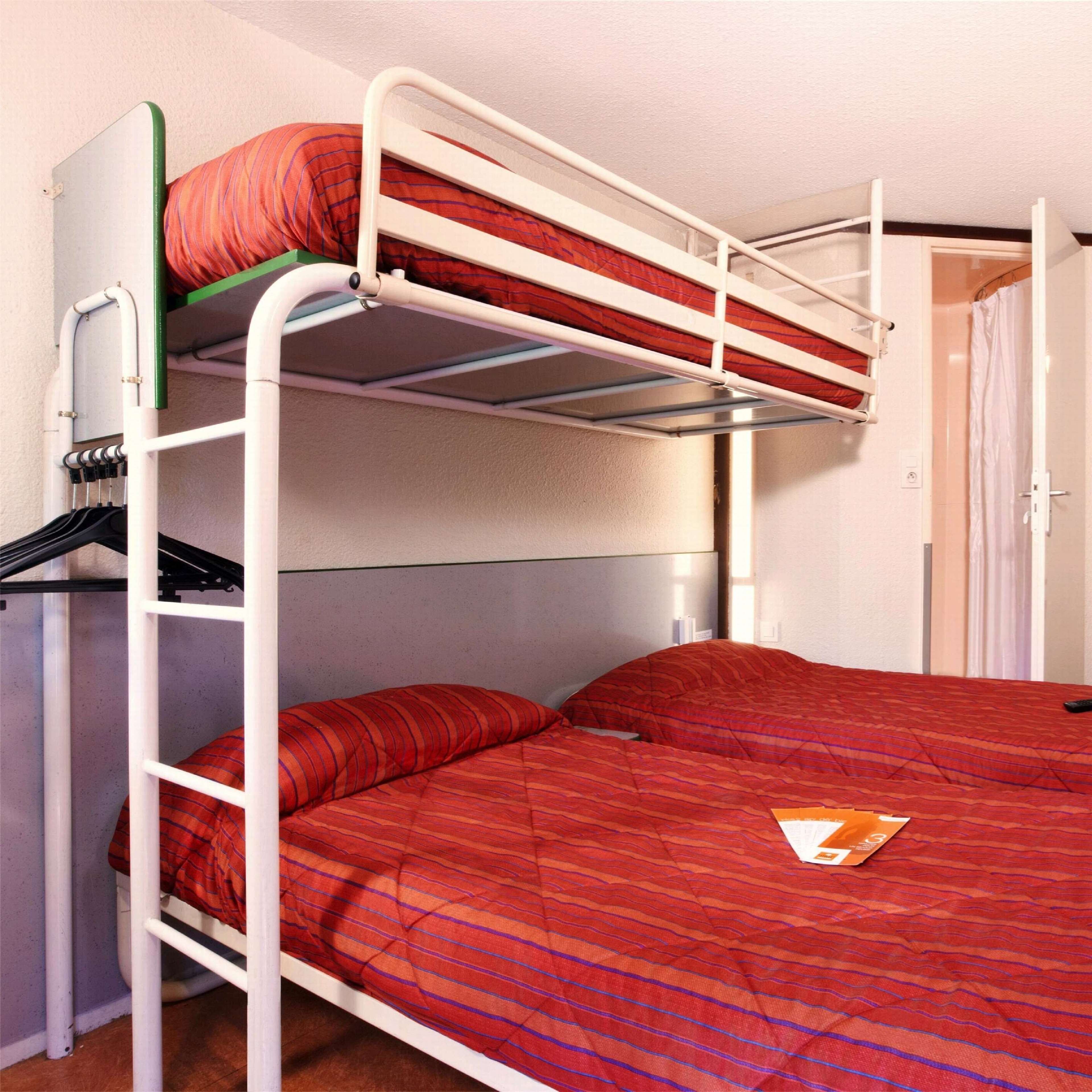 A bunk bed with a red striped mattress and pillows, a white metal frame, and a small shelf with a black bag.