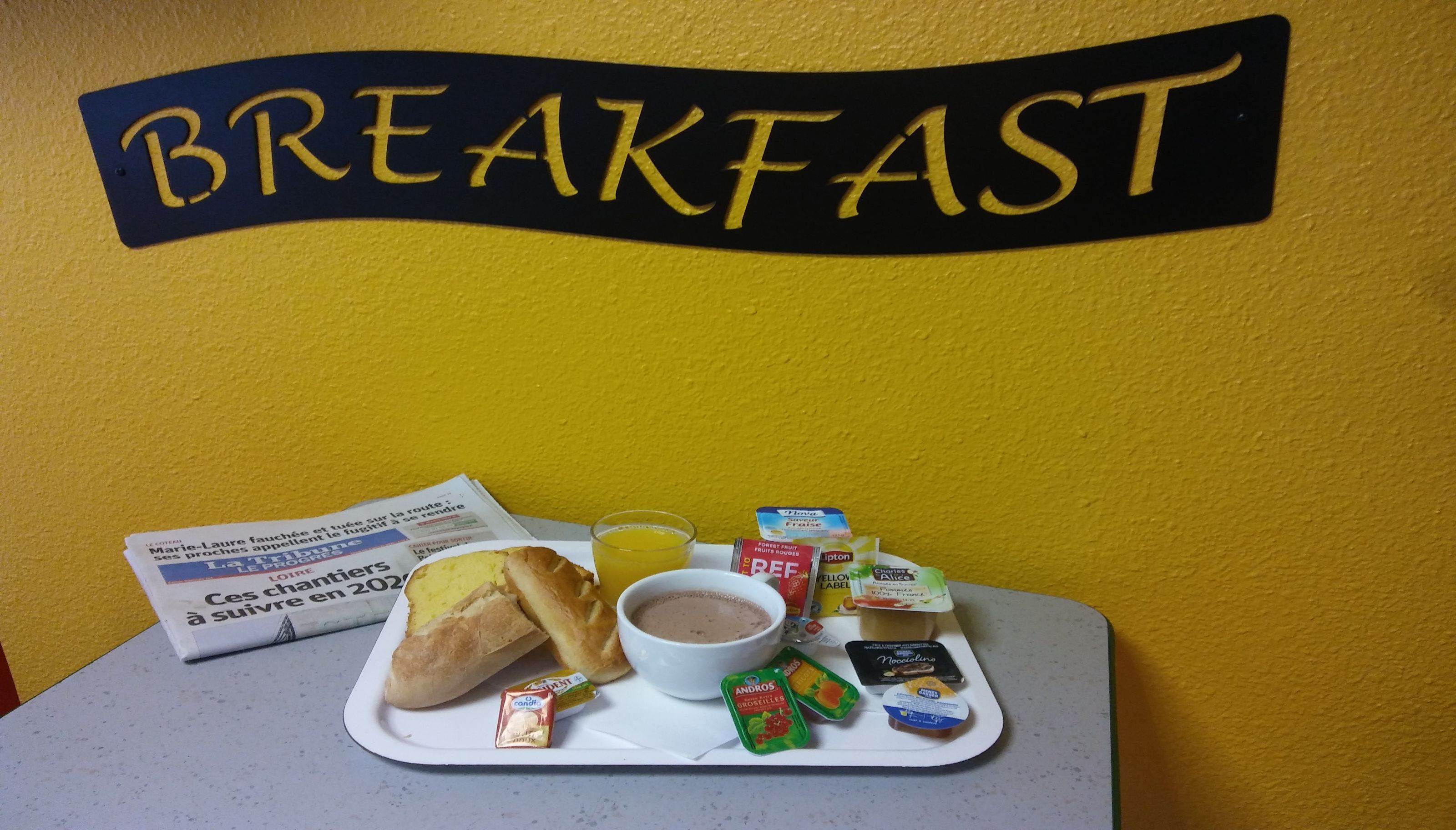 A breakfast tray with bread, a bowl of soup, juice, and various spreads on a table against a yellow wall with a 'BREAKFAST' sign.