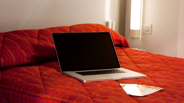 Un ordinateur portable posé sur un lit, recouvert d’une couverture à motifs rouges et agrémenté d’un oreiller blanc.