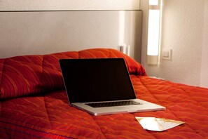 A laptop on a bed with a red patterned bedspread and a white pillow.