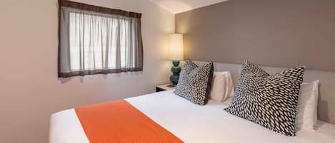 Quarto de hotel com cama, janela com cortinas, abajur e almofadas estampadas.
