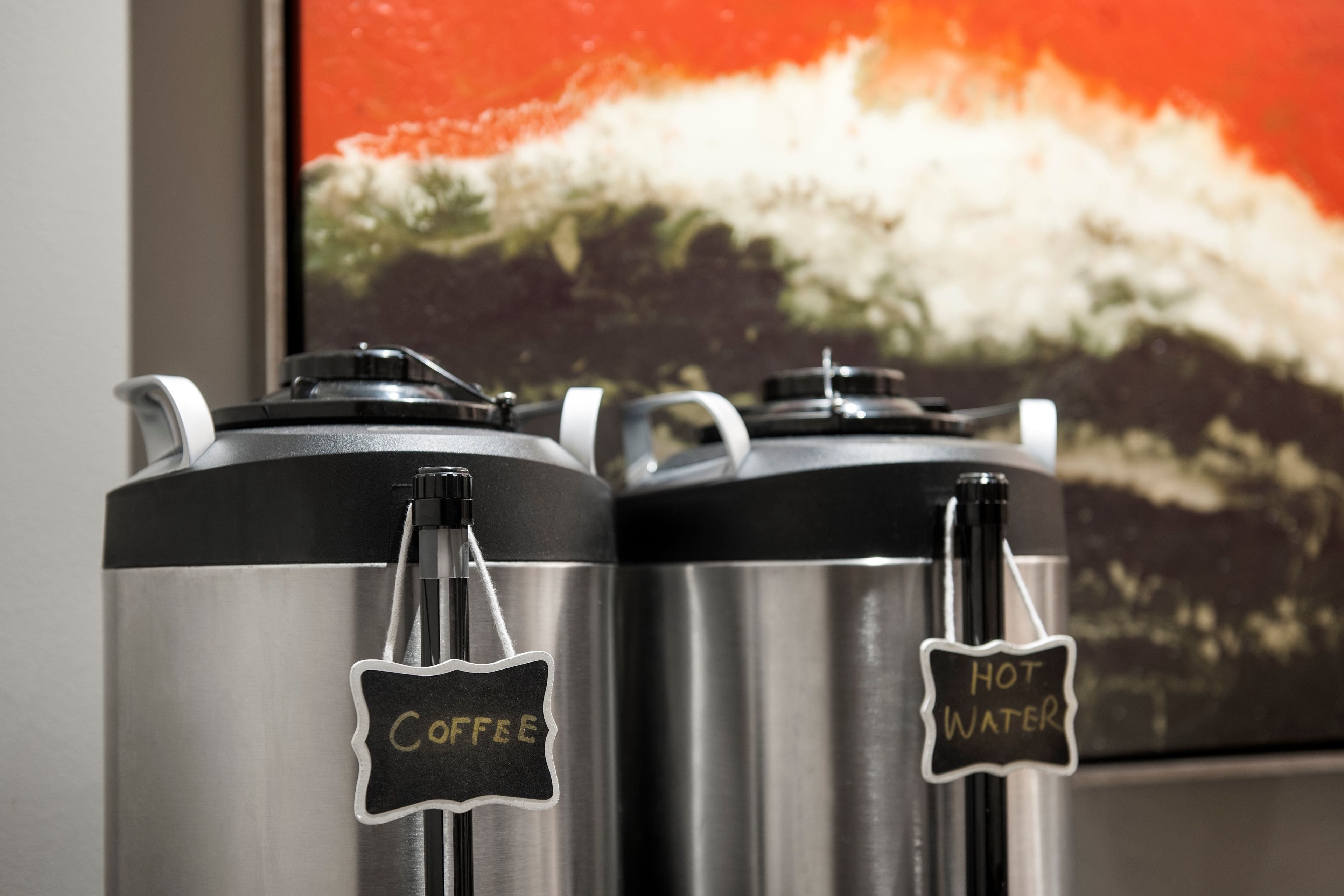 Two stainless steel dispensers labeled "Coffee" and "Hot Water" with a wave painting in the background.