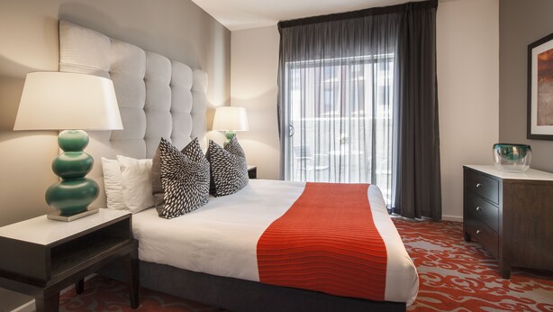 Habitación de hotel con una cama grande, dos mesitas de noche, un tocador y un armario, además de un ventanal con cortinas.