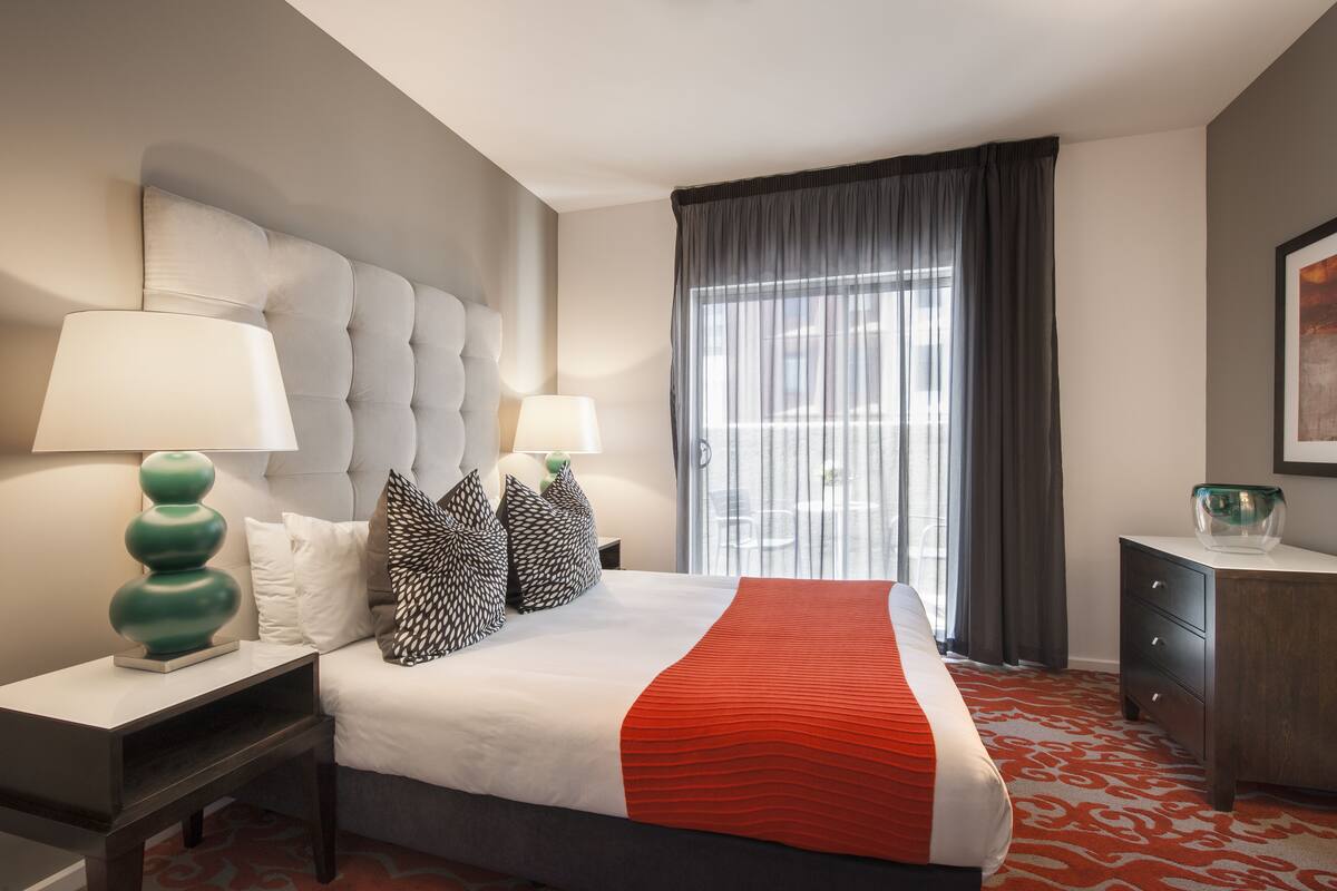 Habitación de hotel con una cama grande, dos mesitas de noche, un tocador y un armario, además de un ventanal con cortinas.