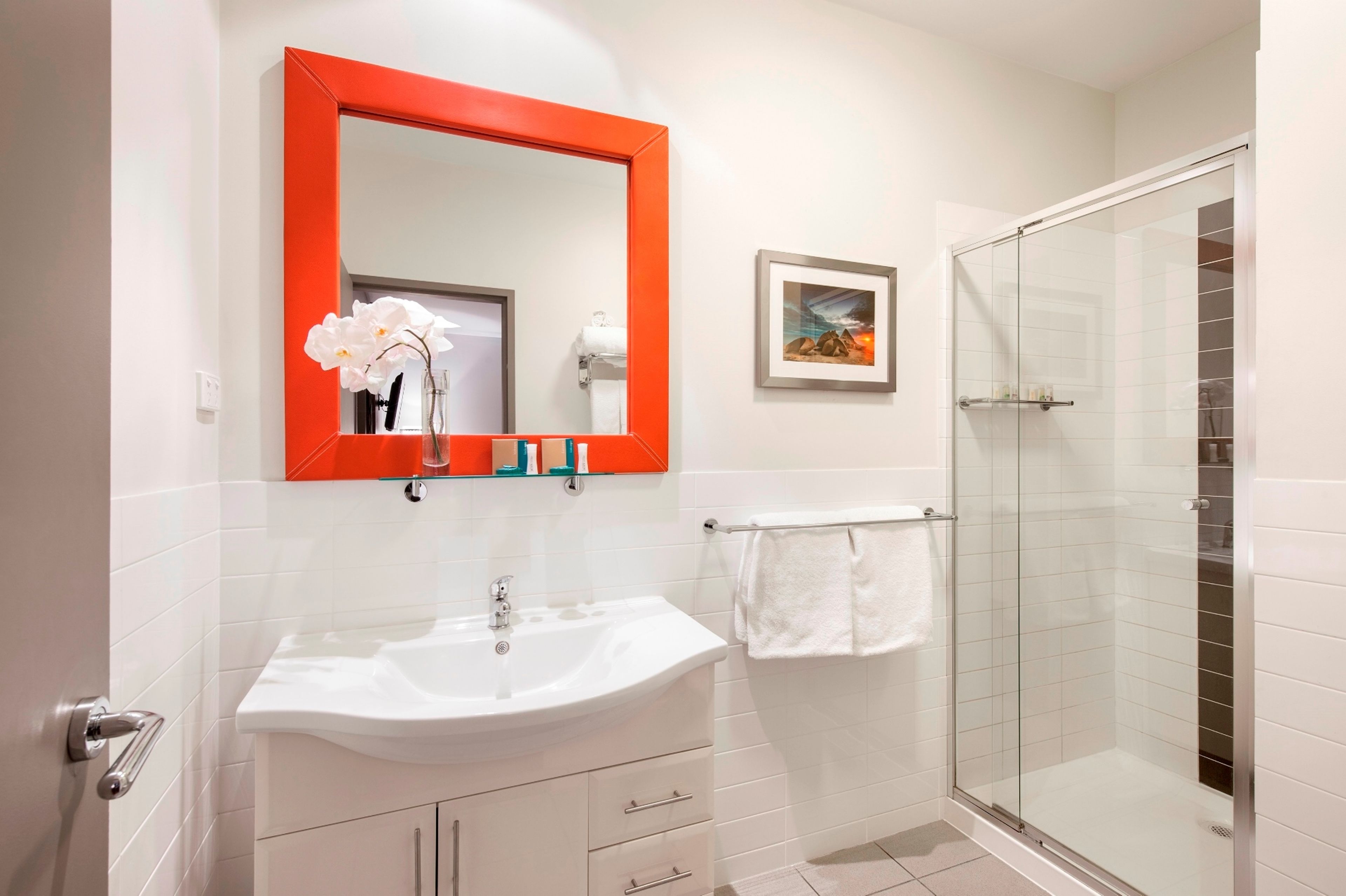 A bathroom with a white sink, a red-framed mirror, a glass shower enclosure, and a framed picture on the wall.