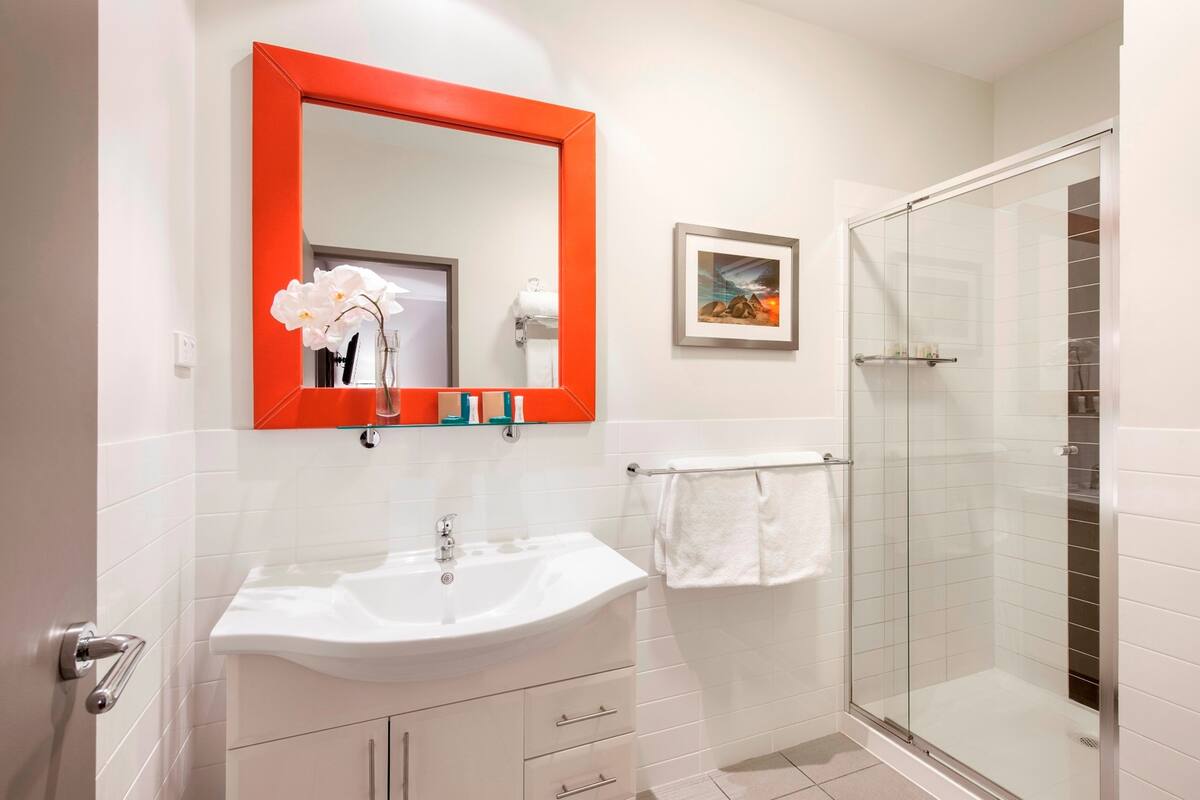 Un baño con un lavamanos blanco, un espejo con marco rojo, una cabina de ducha de vidrio y un cuadro en la pared.