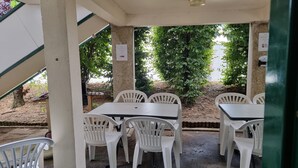 Un patio al aire libre con mesas y sillas de plástico blanco, rodeado de follaje y un piso de concreto.