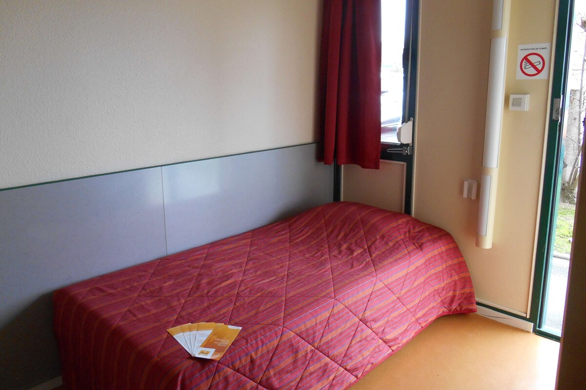 Ein Einzelbett mit roter Bettdecke, ein Fenster mit rotem Vorhang und ein Rauchverbot-Schild an der Wand.