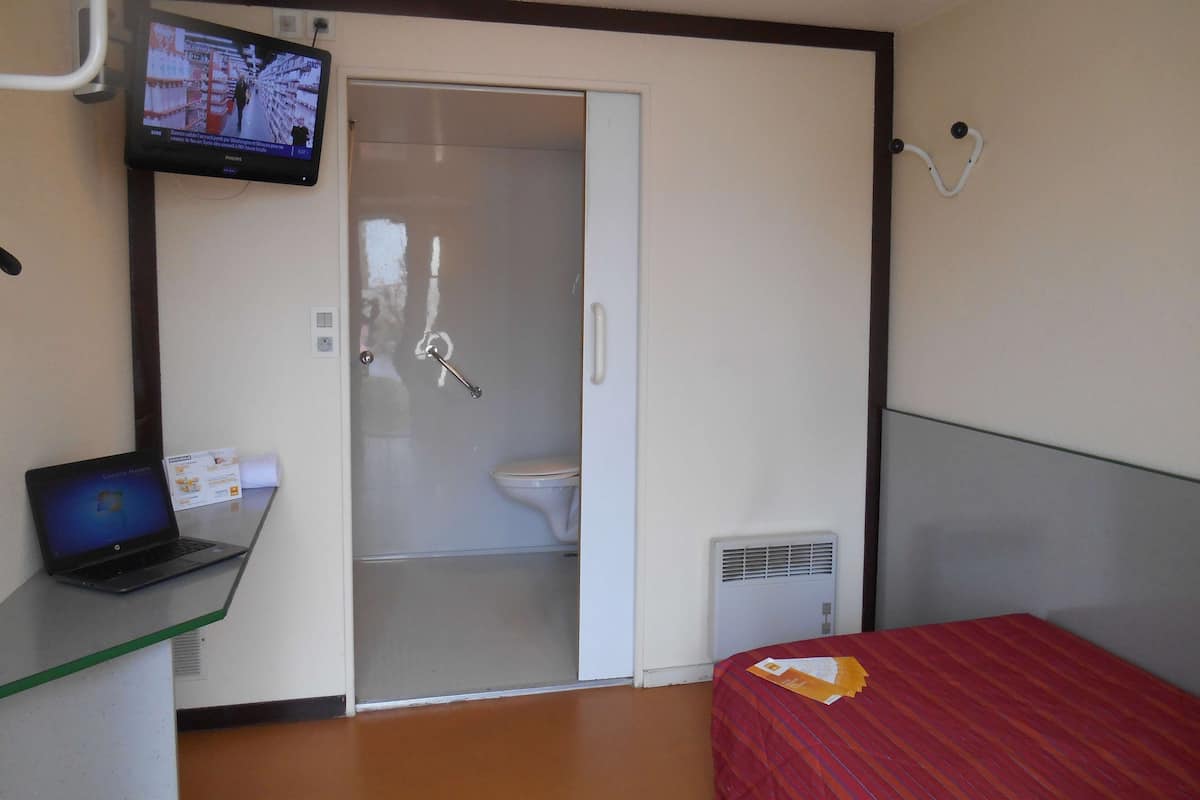Habitación de hotel con cama, escritorio con una computadora portátil, televisor montado en la pared, inodoro y zona de ducha.
