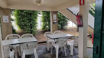 Un área de estar al aire libre con sillas y mesas de plástico blanco, rodeada de follaje y un extintor en la pared.