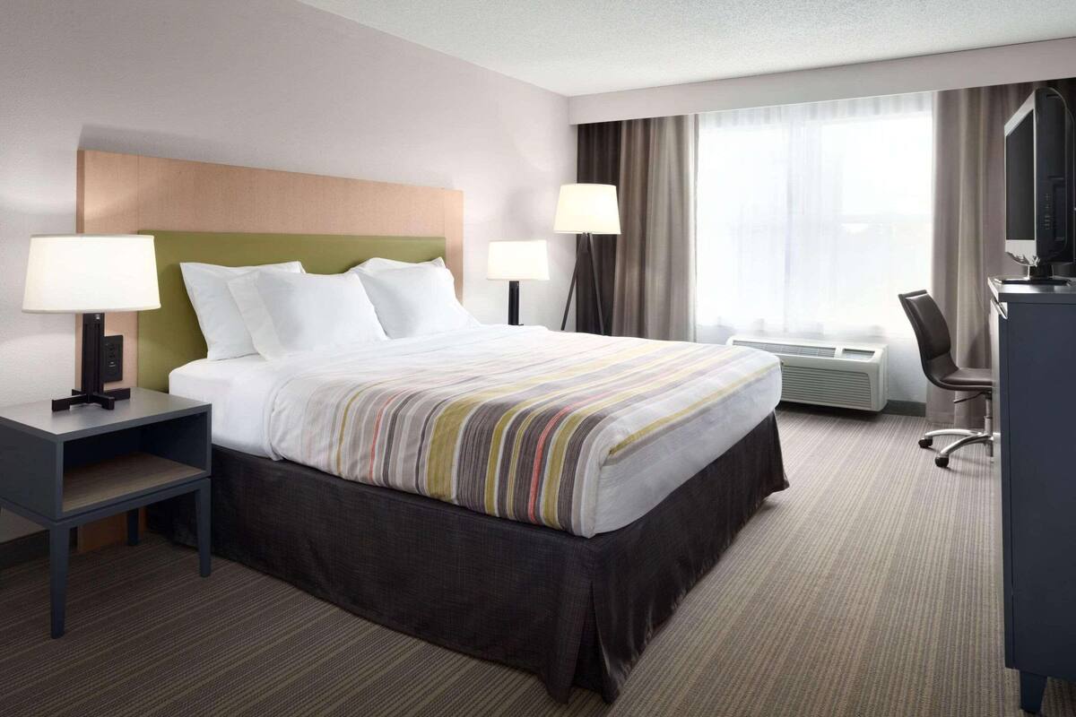 A hotel room with a large bed, two bedside tables with lamps, a desk, a chair, and a large window with curtains.
