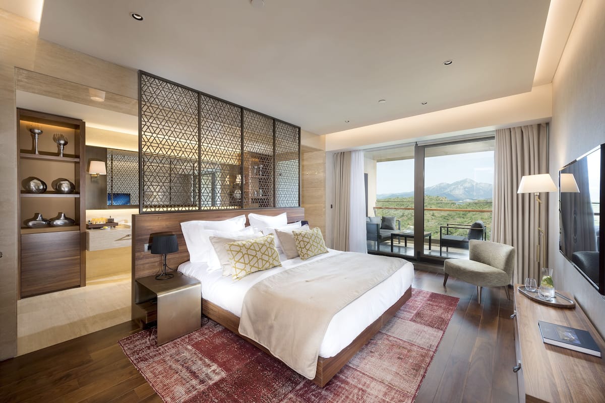 A modern hotel room with a large bed, a TV, a seating area, and a balcony with a mountain view.