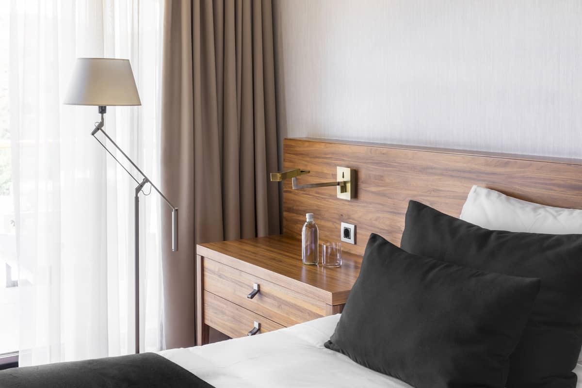 A hotel room with a wooden headboard, a bedside table with a lamp, a bottle, and glasses, and a bed with pillows.