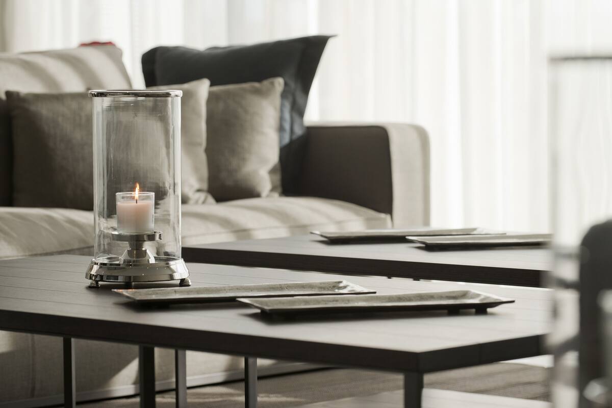 A modern living room with a glass candle holder on a dark wooden table, a sofa with striped cushions, and a window with sheer curtains.