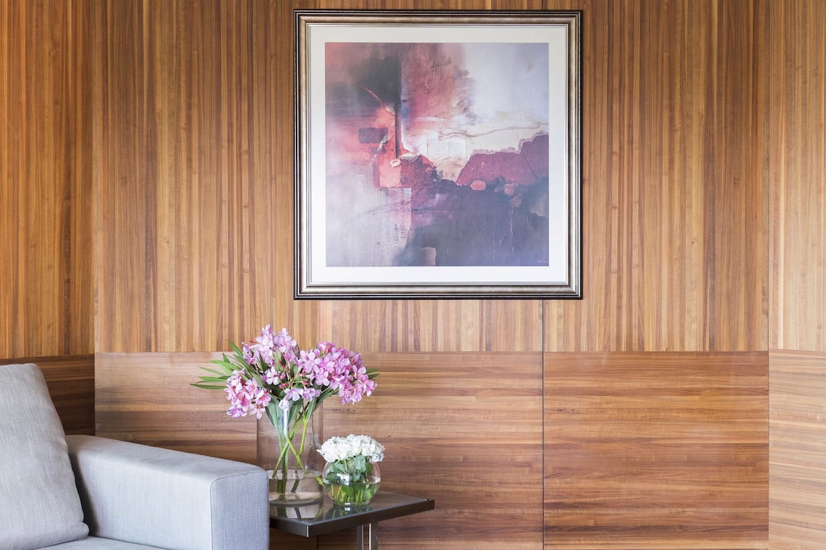 A modern living room with a wooden panel wall, a framed abstract painting, a glass coffee table with flowers, and a grey sofa.