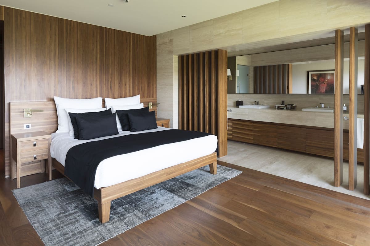 A modern bedroom with a wooden bed, a nightstand, a large mirror, and a bathroom with a wooden vanity and sink.