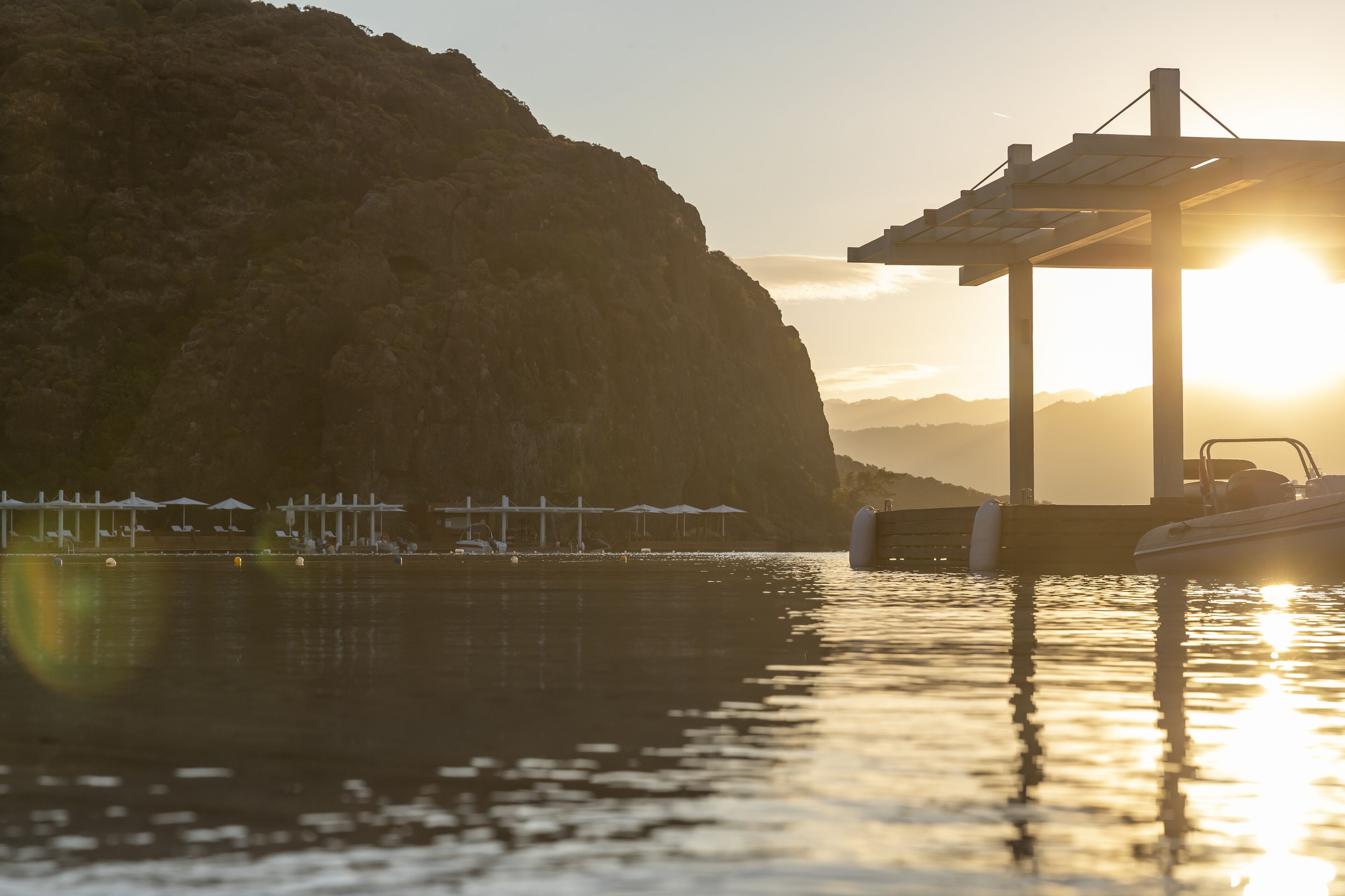 A waterfront scene with a pier, boats, and a mountainous backdrop during sunset.