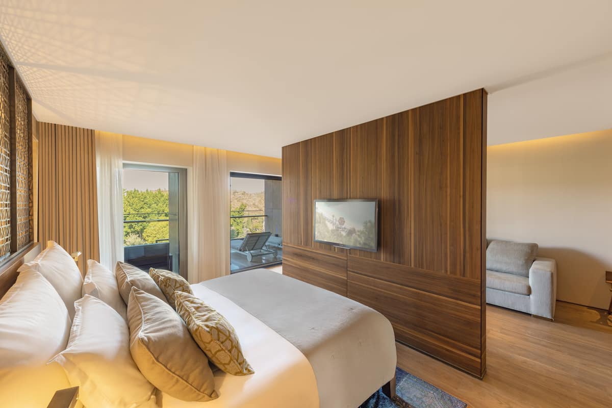 A modern hotel room with a large bed, a TV mounted on the wall, a balcony with a view, and a seating area with a sofa.