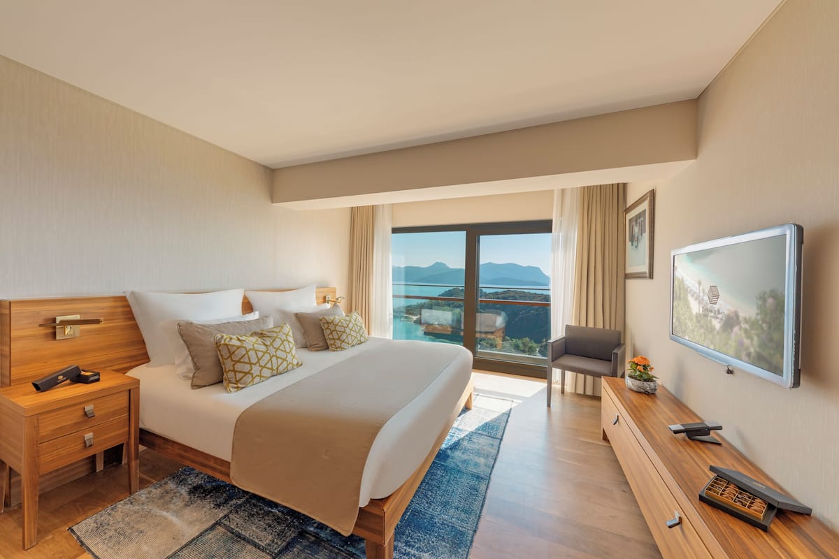A hotel room with a large bed, a wooden nightstand, a desk, a flat-screen TV, and a view of the mountains through the window.