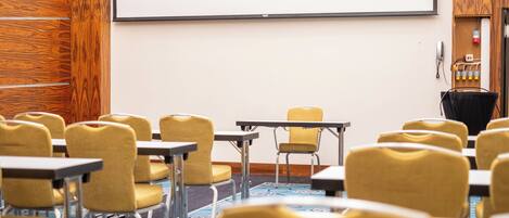 Un salón de conferencias con filas de sillas amarillas y mesas, una pantalla de proyector y un revestimiento de pared de madera.