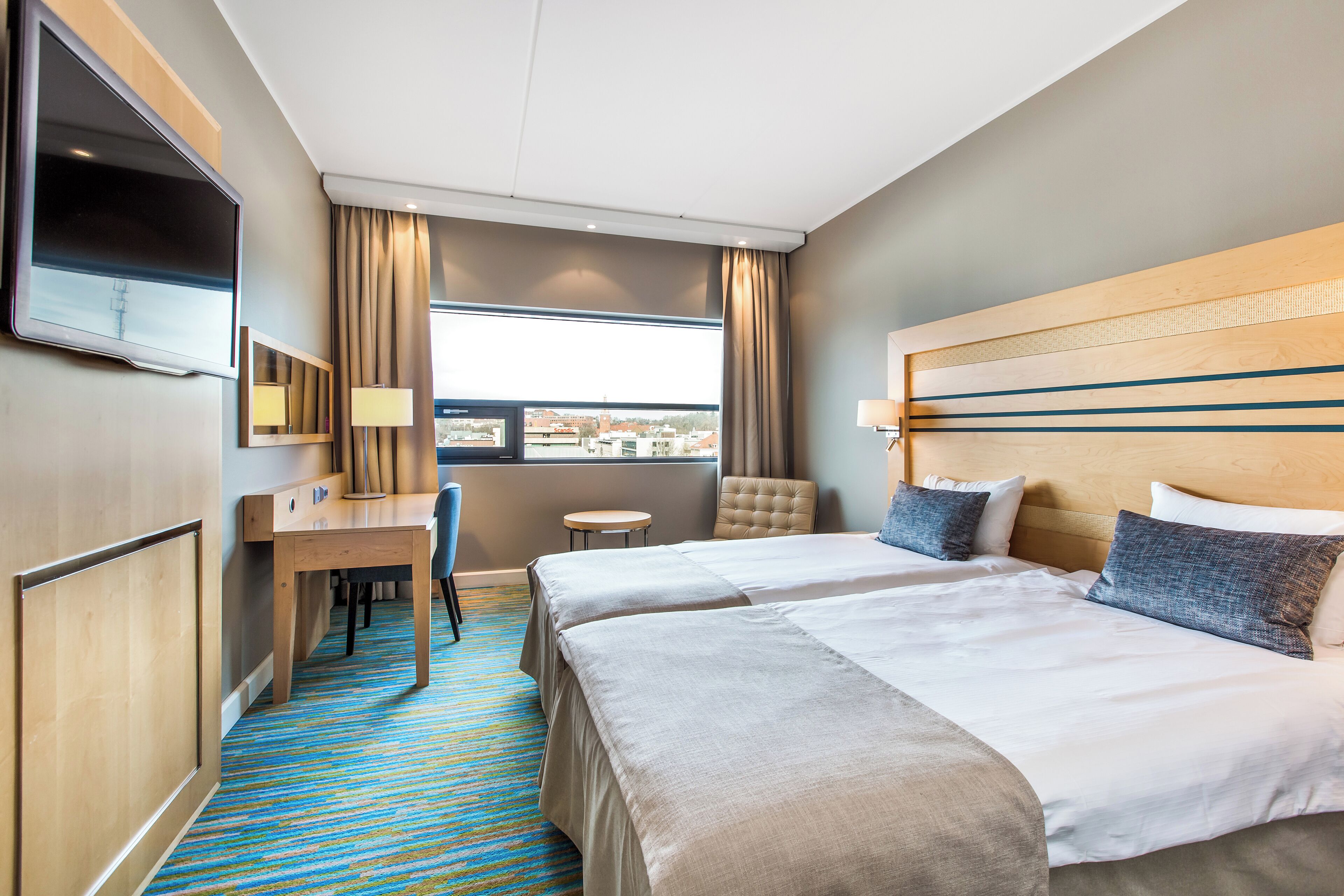 A hotel room with two beds, a desk, a chair, a TV, and a window with curtains.