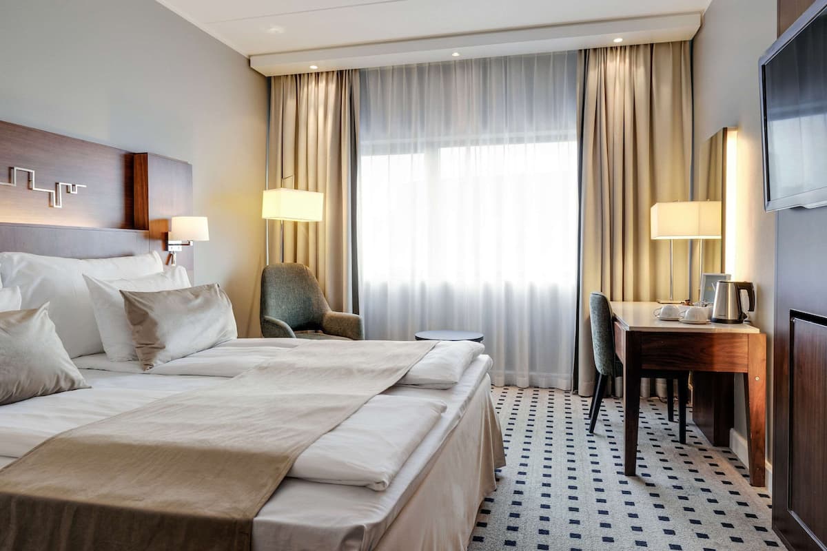 A hotel room with a large bed, two bedside tables, a desk with a chair, a TV, and a window with curtains.