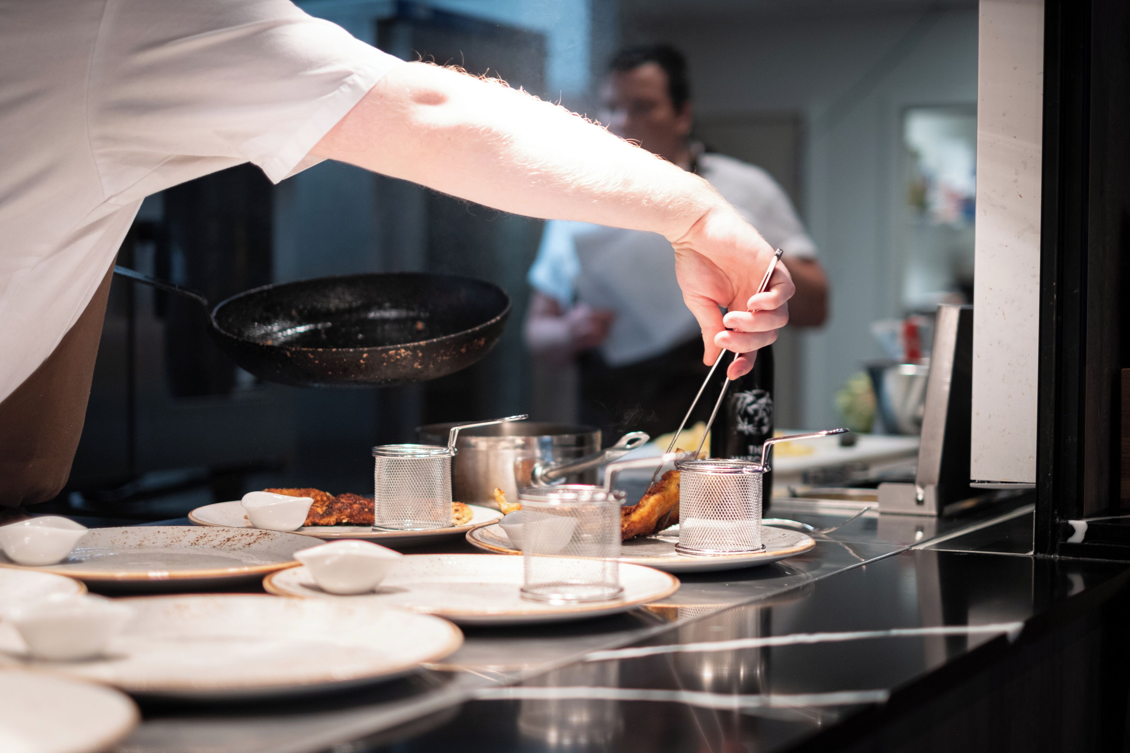 Una mano de chef sosteniendo un utensilio sobre un wok, con varios utensilios de cocina y platos en la encimera.