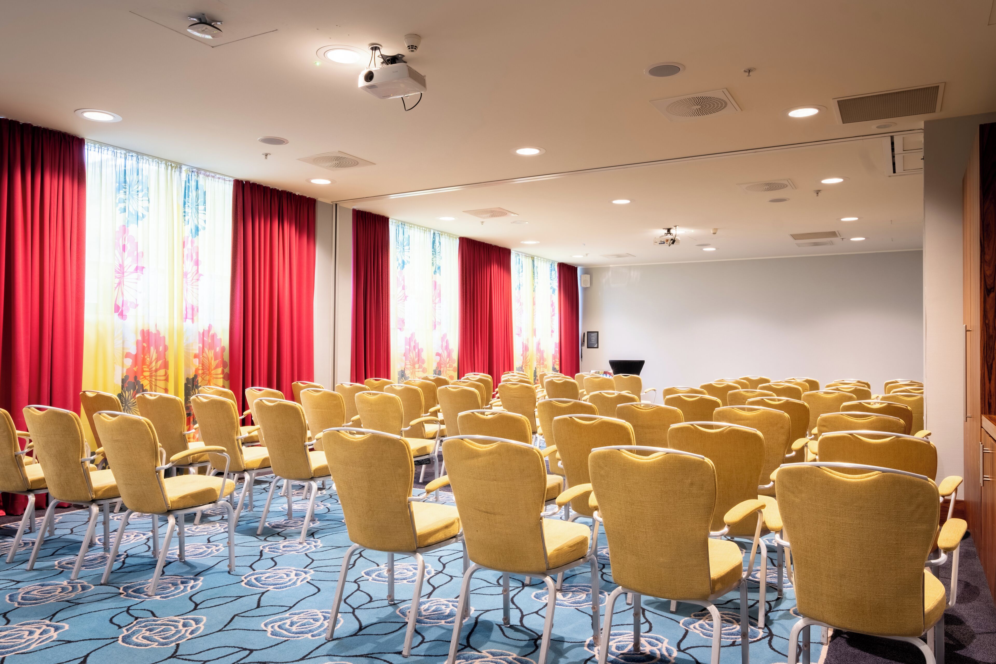 A conference room with rows of yellow chairs, a projector, and red curtains.