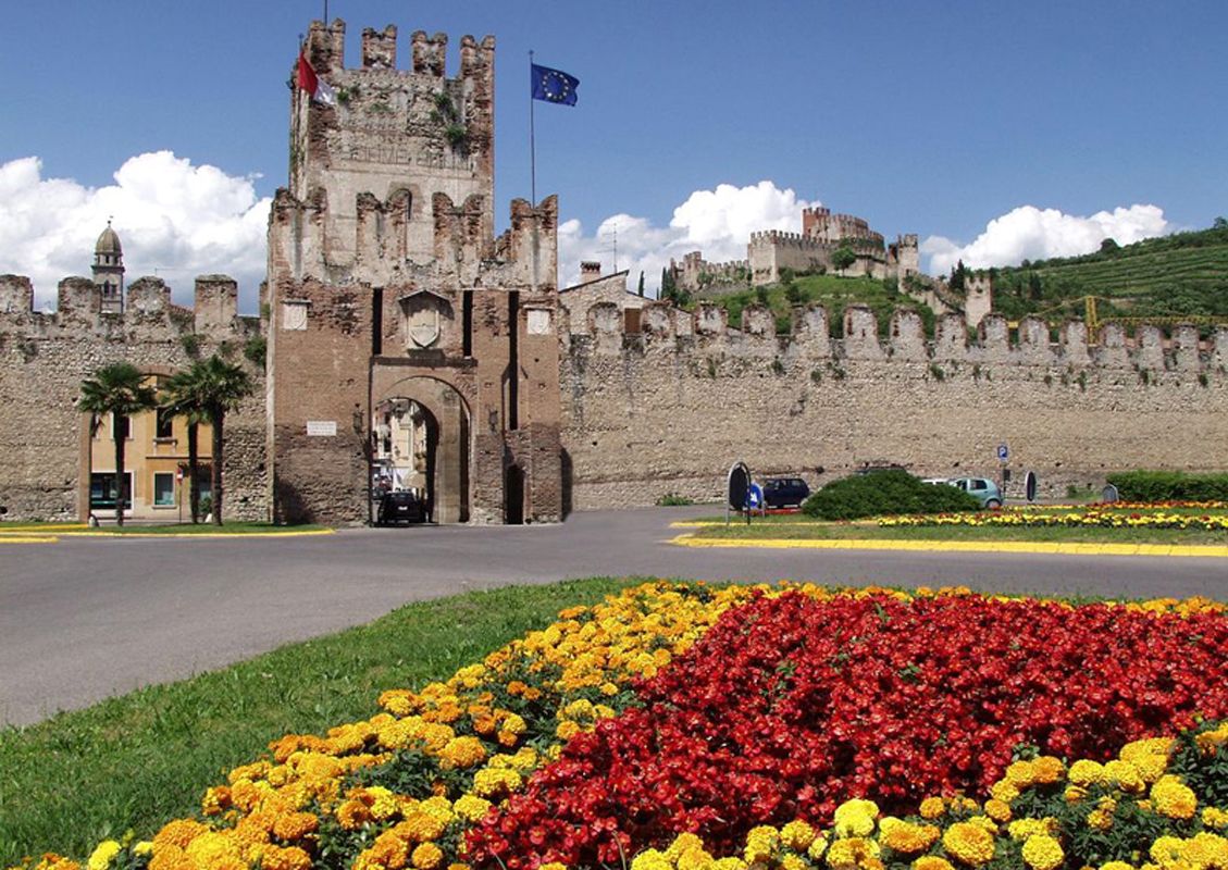 La entrada de un castillo histórico, con una bandera, un jardín de flores y un camino pavimentado.