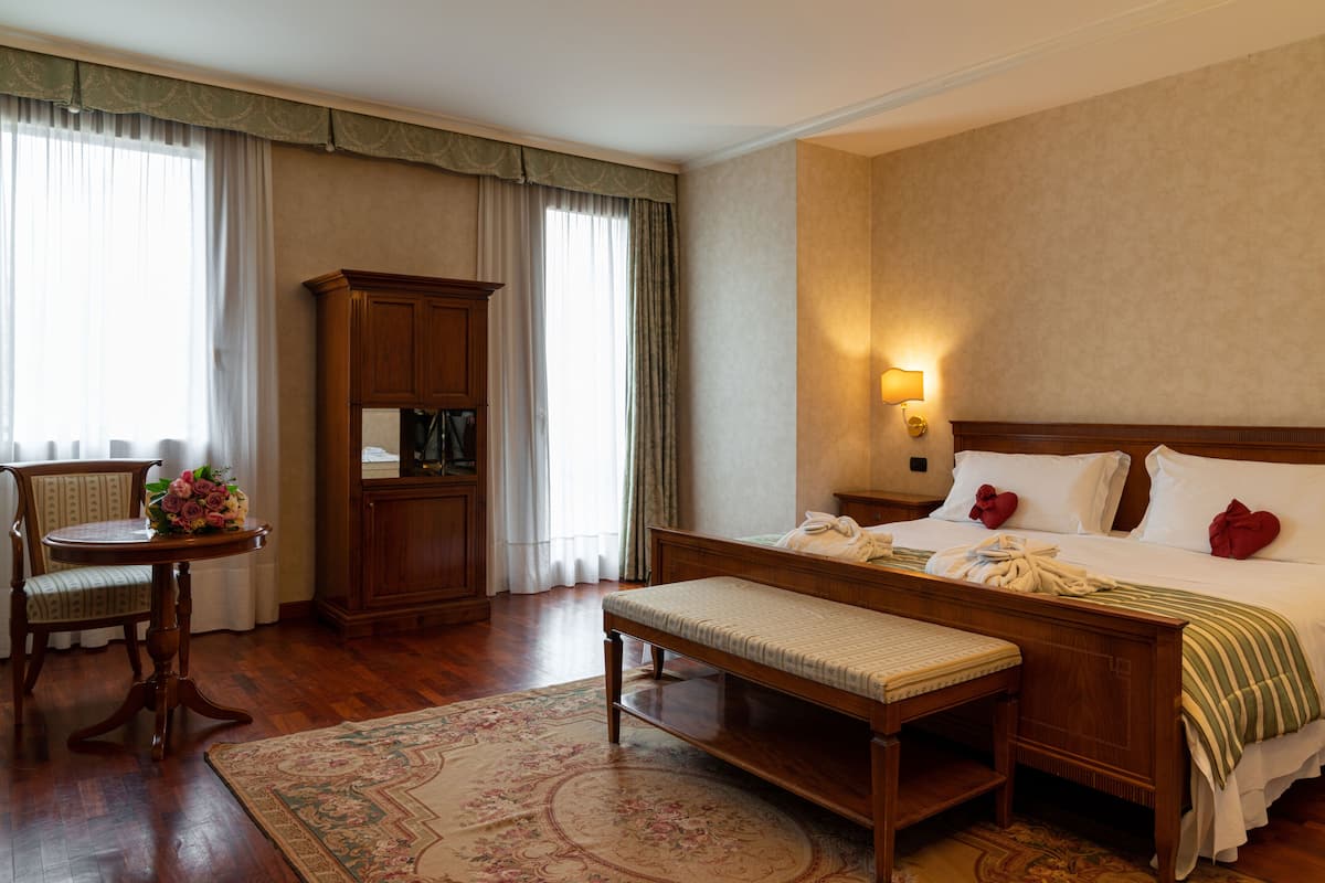 A hotel room with a bed, bedside table, bench, wooden wardrobe, and a desk with a chair and flowers.