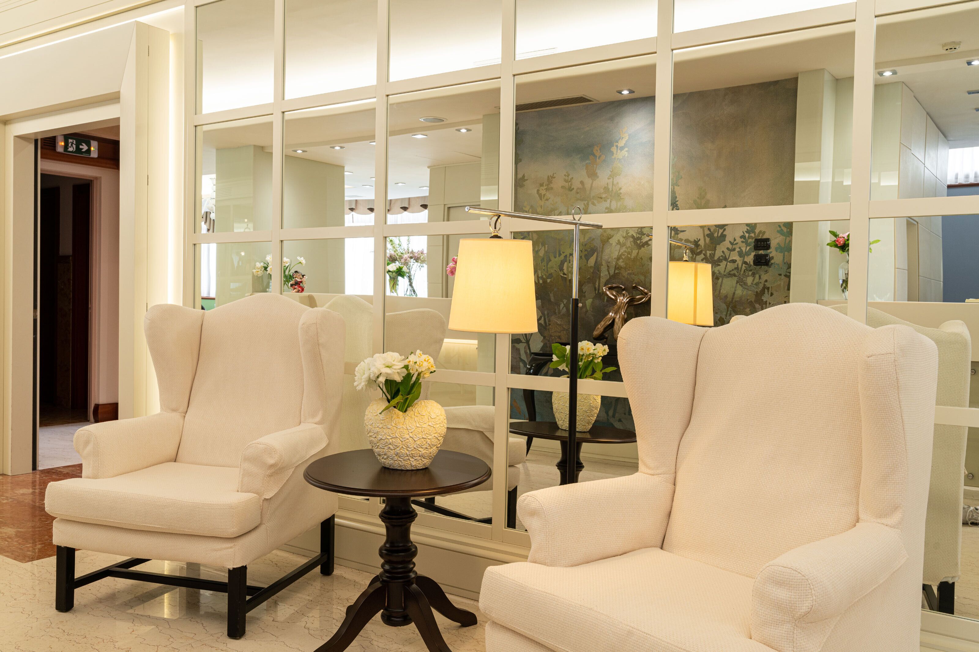 Two white armchairs facing each other with a small round table between them, a vase with flowers on the table, and a large window with reflections of interior decor.